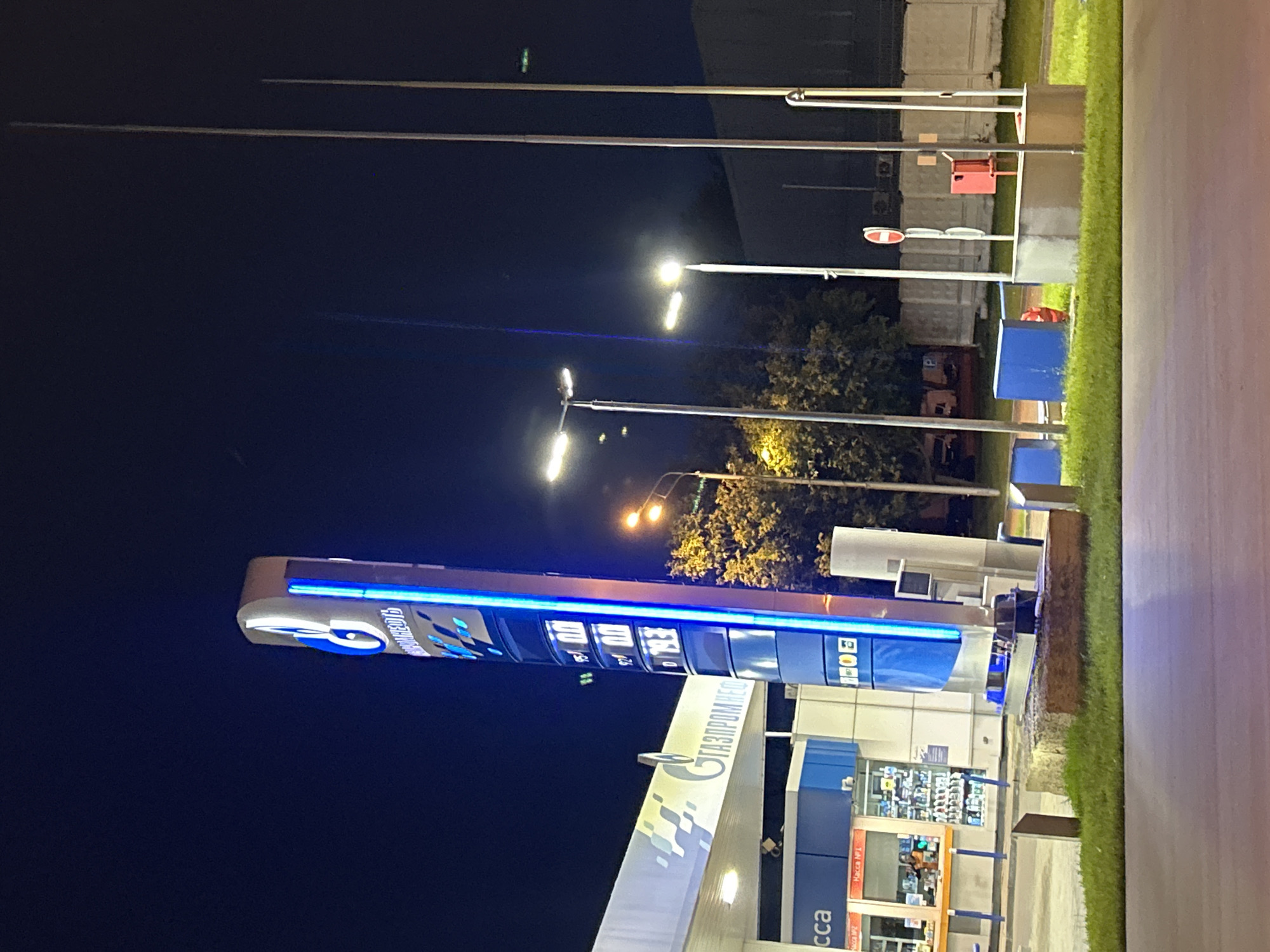 The image depicts a night scene at a Gazpromneft gas station in Moscow, Russia. A tall, illuminated blue sign displays fuel prices, with "95 0.0", "92 0.0", and "D 79.3" visible. To the left of the sign is the covered area of the gas station, with the "GAZPROMNEFT" logo clearly visible in white lettering. A small kiosk with a person visible inside is also present. Several streetlights illuminate the area. In the background, there is a wall, trees, and other structures. The overall scene is indicative of a typical urban gas station environment at night.