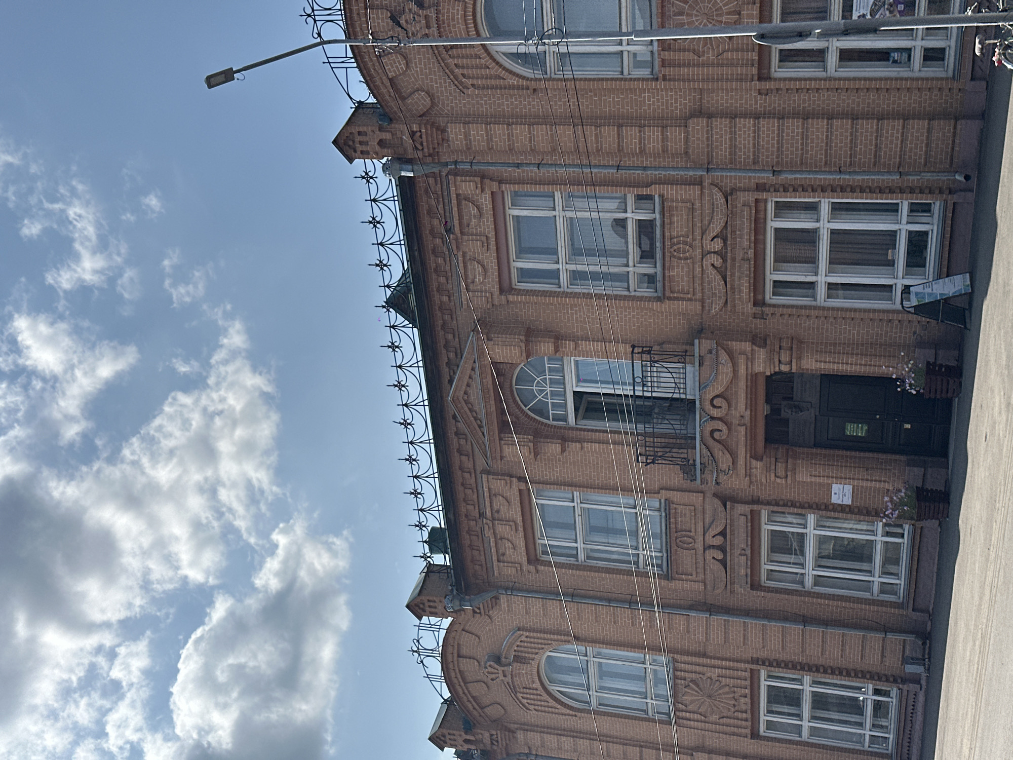 This image shows a brick building in Biysk, Russia. The building has a light pinkish-orange brick facade with ornate architectural details, including arched windows with decorative grilles, a balcony with an open door above it, and a decorative metal railing along the roofline. Several windows on the facade are visible, some of which are large and multi-paned. A modern street lamp is situated to the right of the building, extending upwards into a blue sky with scattered clouds. Power lines are strung across the facade. At the base of the building, there is an entrance with a dark-colored door, flanked by flower planters. A-frame sign stands near the entrance. A bicycle is partially visible at the bottom right corner of the image. The overall scene depicts a street view of a historic building under a bright sky.