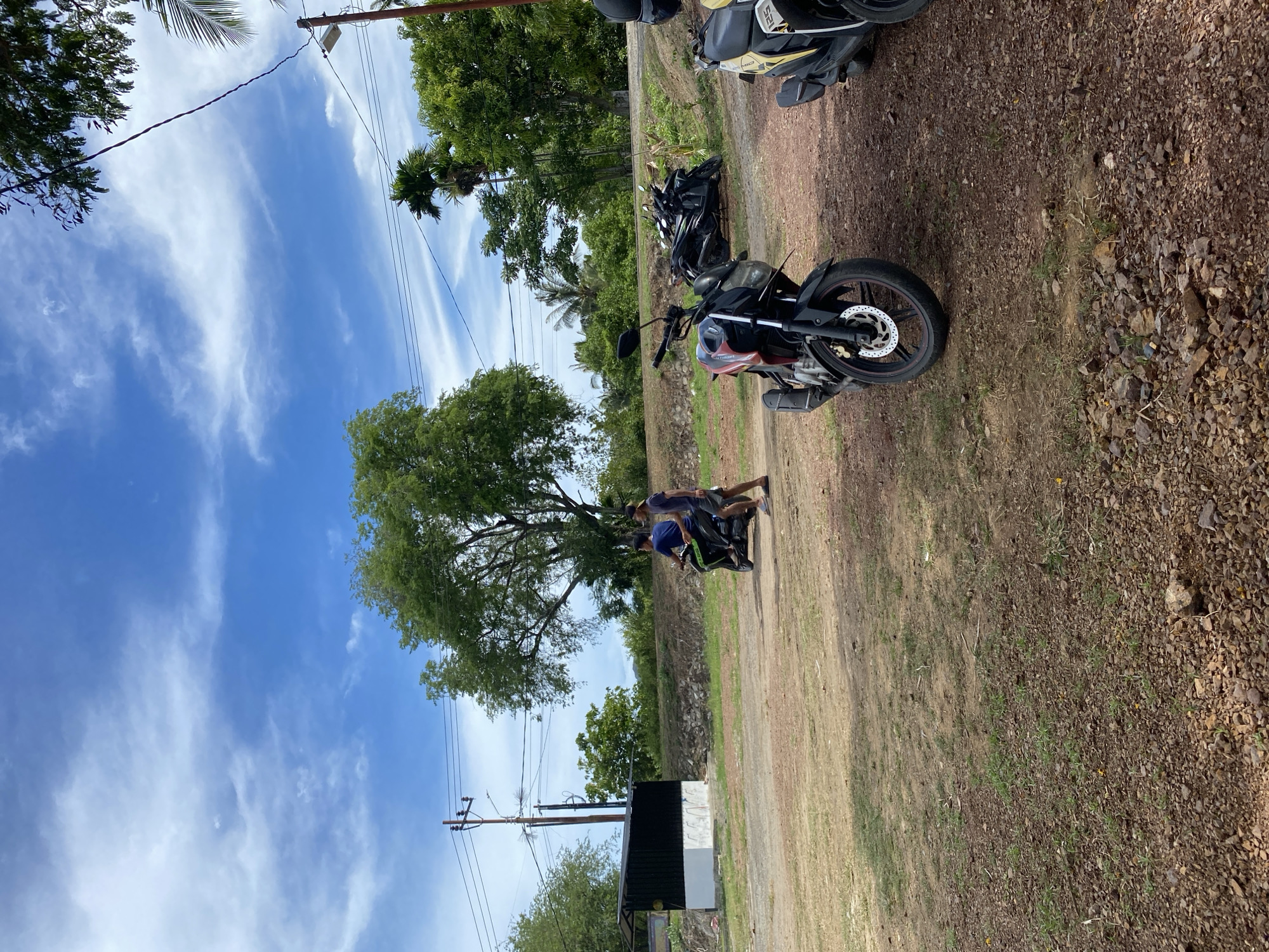 The image depicts an outdoor scene with a dry, dusty ground. In the foreground, two motorcycles are parked. One is a black and red motorcycle with a visible front wheel, disk brake, and red rim accents. Partially visible to its right is a yellow and black scooter. Further back, a green motorcycle is being ridden by one person, with another person walking alongside it. Behind them, a black building with a white base is situated. The background features a line of trees and lush green vegetation. The sky is a bright blue with scattered white clouds, and utility poles with electrical wires are visible in the distance. The scene appears to be in a rural or semi-rural area, likely within the city of Aneuk Paya, Indonesia, given the context.