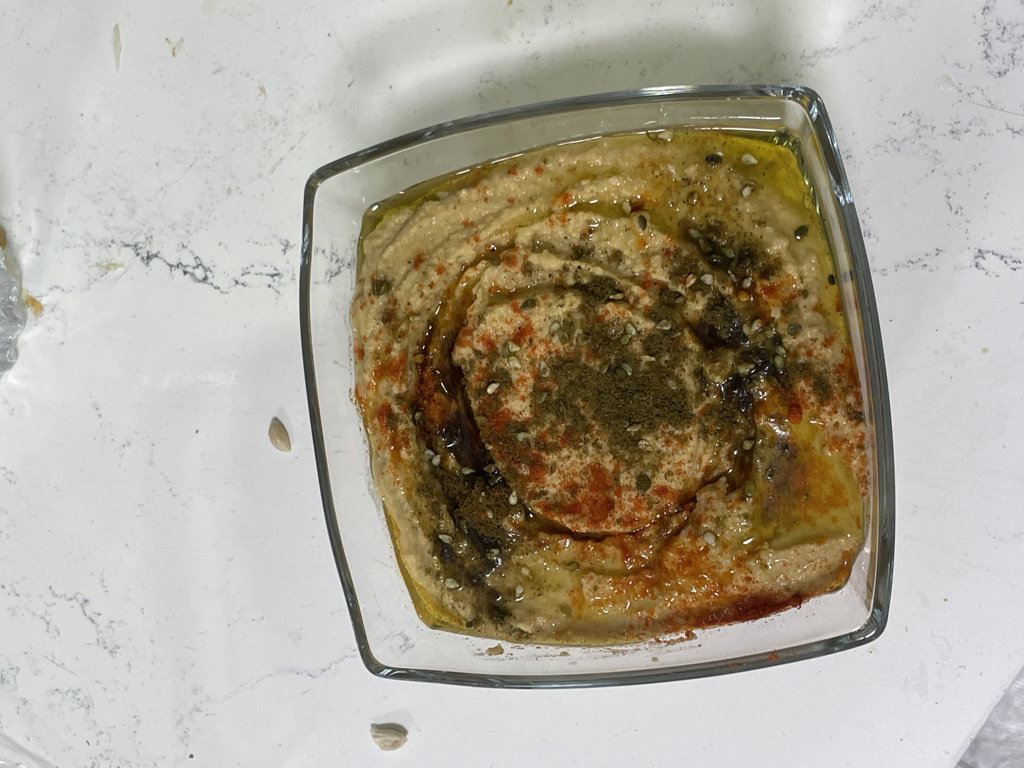 A close-up, overhead shot showcases a square glass bowl filled with hummus. The hummus has a swirl pattern, drizzled with olive oil and topped with a sprinkle of paprika and what appears to be za'atar. Several whole chickpeas are scattered on the white, speckled countertop surrounding the bowl. The scene is set on a countertop in Raanana, Israel.