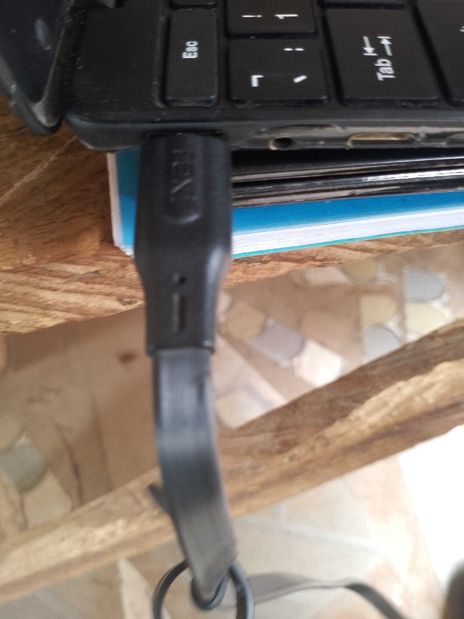A close-up, slightly tilted, high-angle shot shows a black USB cable plugged into a laptop. The cable has a black connector with the word "REX" visible in white lettering. The connector is plugged into a port on the side of a laptop, the edge of which is visible at the top of the frame. The laptop keyboard is partially visible, with keys labeled "Esc" and "Tab" clearly seen. A stack of books with blue and white covers is positioned beneath the laptop and behind the USB cable. The background is blurred and appears to be an indoor setting with wooden furniture and possibly a tiled floor. The lighting suggests it is daytime.