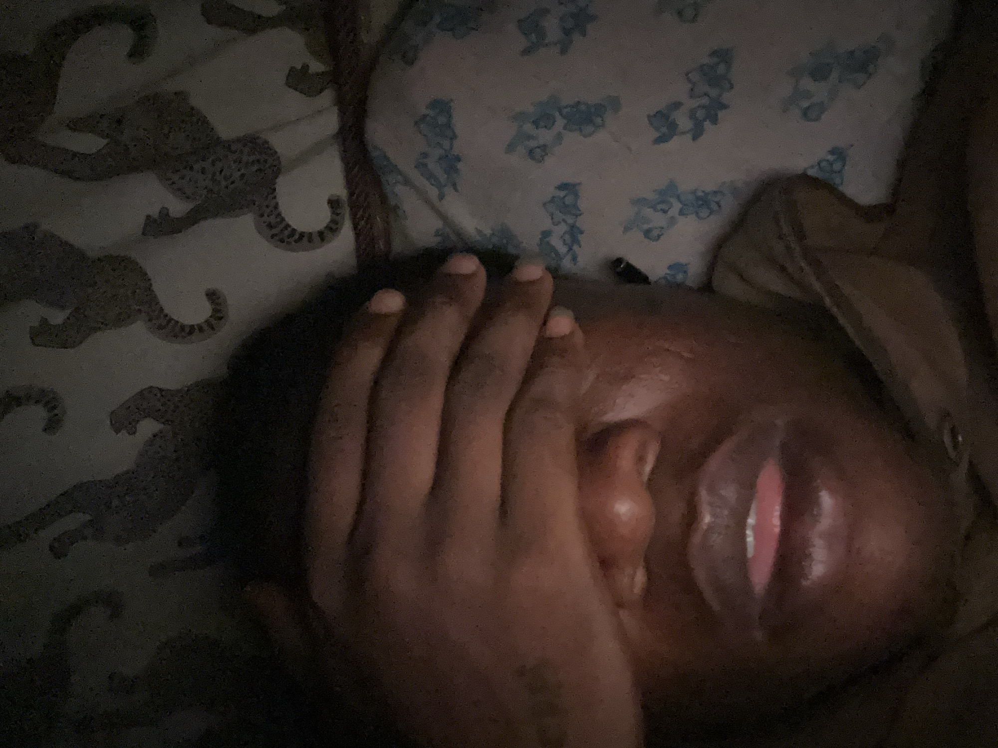A person with dark skin is lying down with their hand covering their face. The person's lips are visible. The person is lying on a bed with two different patterns on the pillow and the comforter. The pillow has a blue and white floral pattern. The comforter has a grey animal print pattern. The scene is dimly lit.