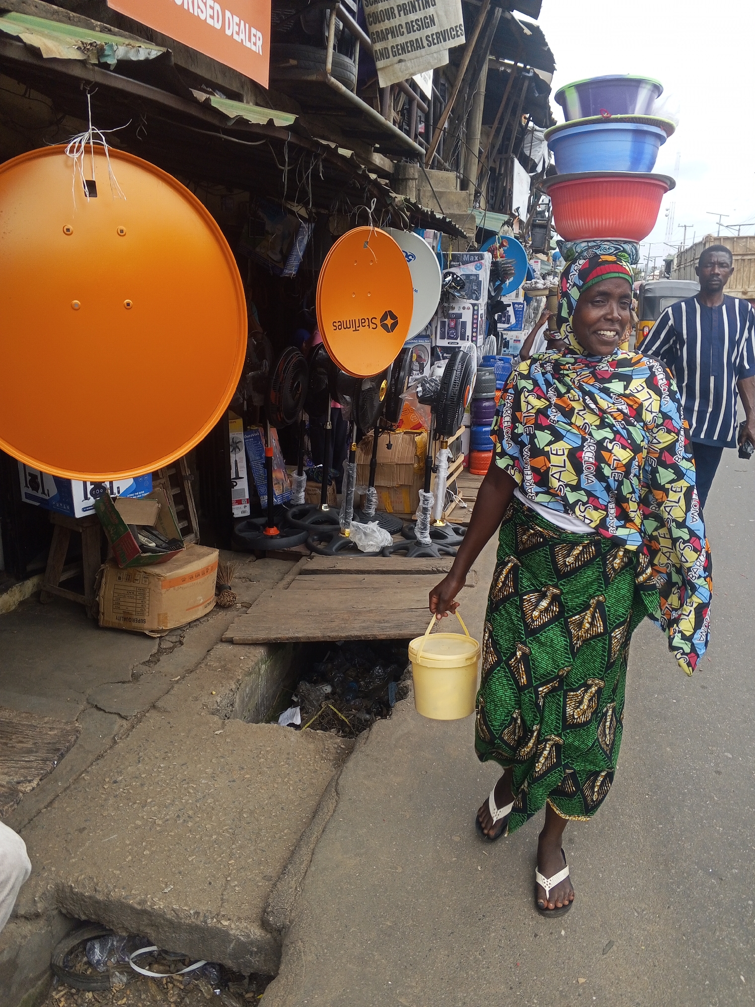 In Okene, Nigeria, a woman walks along a street lined with market stalls. She carries a yellow bucket in her right hand and has a stack of colorful plastic basins balanced on her head. She is smiling and looking towards the camera. She wears a headscarf and a patterned skirt and top. Behind her, a man in a striped shirt walks away from the camera. The market stalls display various items, including satellite dishes, fans, and electronics. The ground is paved with concrete and there is a drainage ditch filled with trash. The overall scene depicts a bustling street market environment.