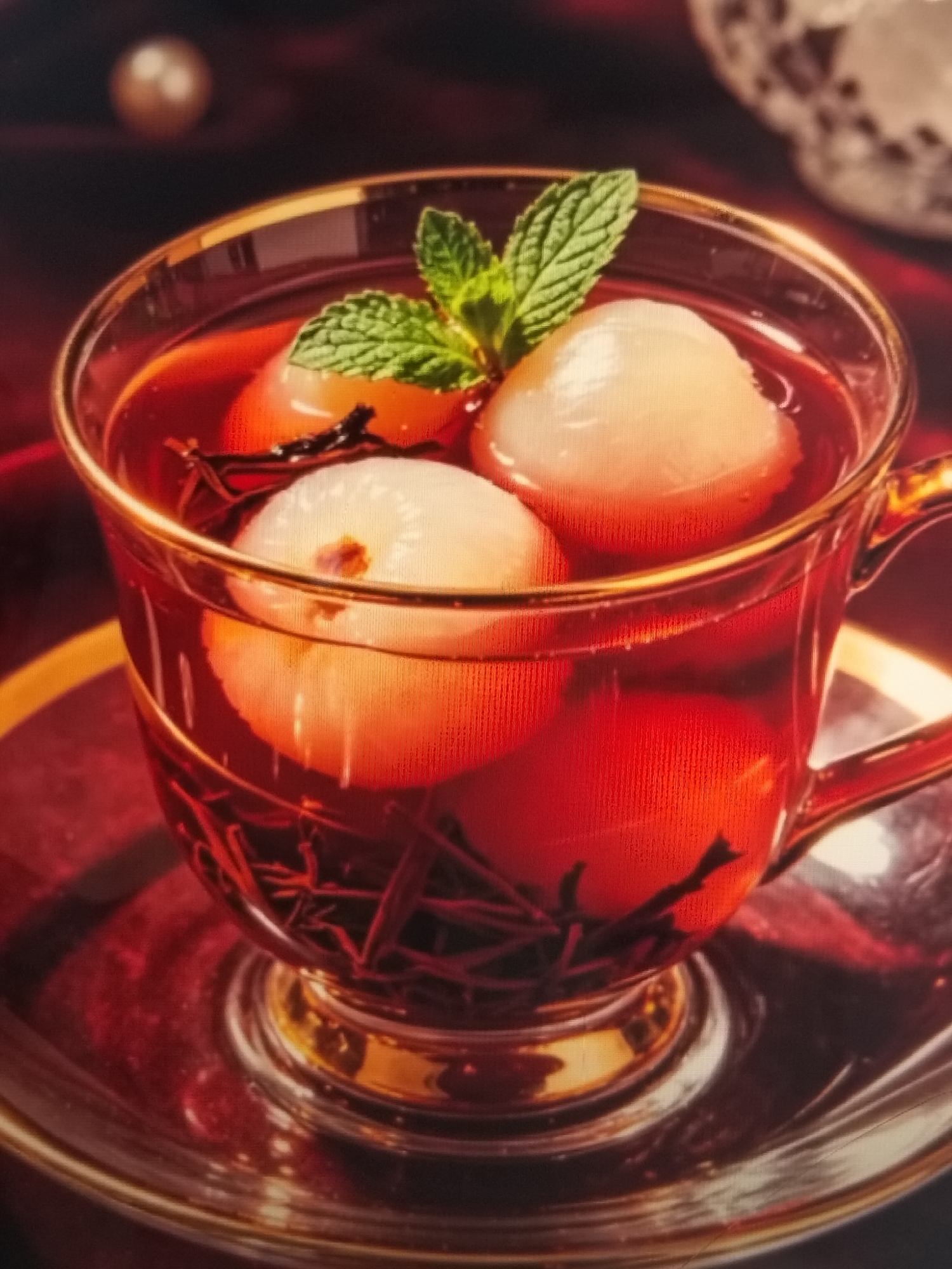 The image shows a close-up of a clear glass teacup filled with a reddish-brown liquid and several white, round fruits, likely lychees. A sprig of green mint is placed on top of the fruits. Dark, thin strands, possibly tea leaves or spices, are visible within the liquid. The teacup has a gold rim and a gold-banded handle, and it rests on a matching saucer. The background is blurred, with hints of dark red fabric and a faint, out-of-focus object resembling a pearl in the upper left. There is no discernible information about the city of Payagaboh, Indonesia, or any people in the image.