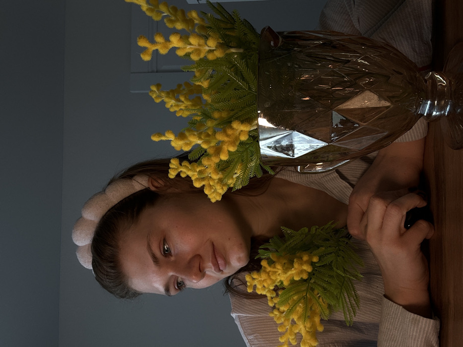 An indoor scene in Nikol'skii, Russia, depicts a female subject seated at a dark wooden table. She wears a light-colored, padded headband and a light-colored, vertically striped shirt. Her hands are clasped on the table surface, and she holds a small cluster of yellow, mimosa-like flowers with green foliage in her left hand. Her head is turned slightly to the left, and her gaze is directed towards the left of the frame. To the right of the subject, a larger arrangement of identical yellow flowers is presented in a clear, faceted glass vase with a footed base. The background consists of a dimly lit, light-colored wall, with what appears to be a door frame visible on the right side.