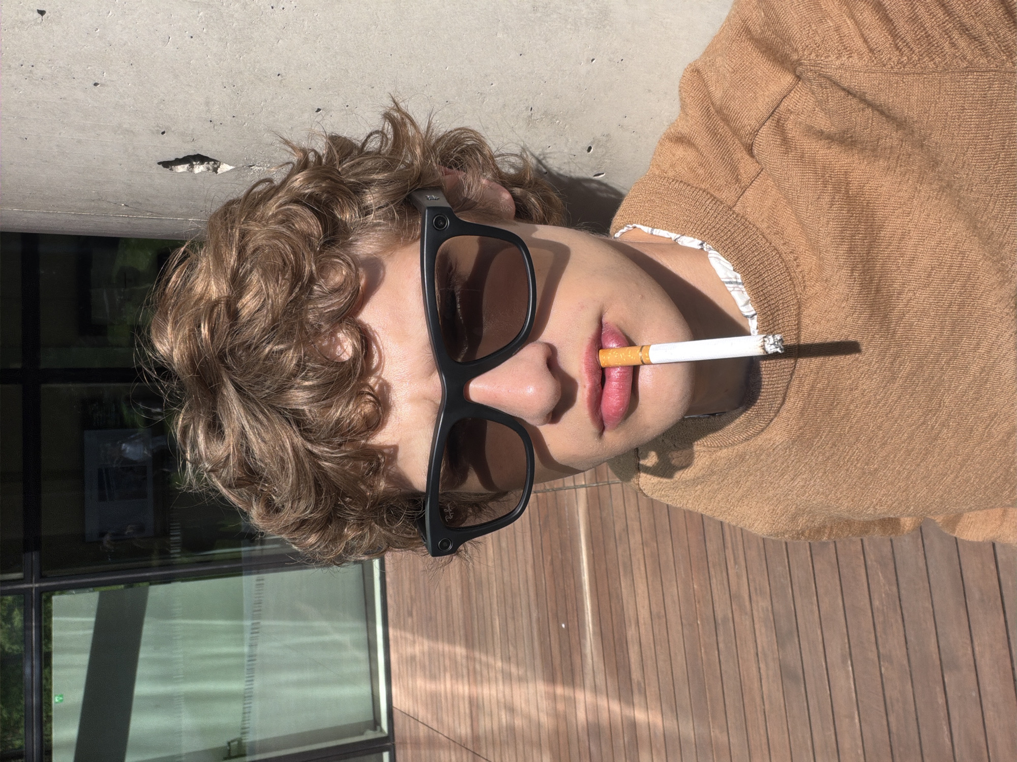 A rotated high-angle shot depicts a person with light brown, curly hair lying down outdoors, under bright sunlight in Mirabella, Italy. The image is rotated 90 degrees clockwise, positioning the person's head on the left and their body extending to the right.
The subject wears black, Wayfarer-style sunglasses, with the "Ray-Ban" logo visible on the right arm. A lit cigarette is held casually between their lips, with a small amount of ash forming at the tip. They are dressed in a light brown or tan knitted sweater, under which a patterned, possibly plaid or checkered, shirt is visible.
Behind the person's head and shoulders is a light gray, textured concrete surface, showing some minor imperfections and a distinct hole. Below their body, a wooden deck with parallel planks is visible, reflecting the strong sunlight. Harsh shadows are cast by the person and the cigarette, indicating a clear, sunny day. Reflections in a window, partially visible in the upper-left of the original image (when oriented upright), suggest an urban or architectural setting nearby. The person's eyes are obscured by the sunglasses, but the pose and cigarette convey a casual, perhaps contemplative or relaxed demeanor.