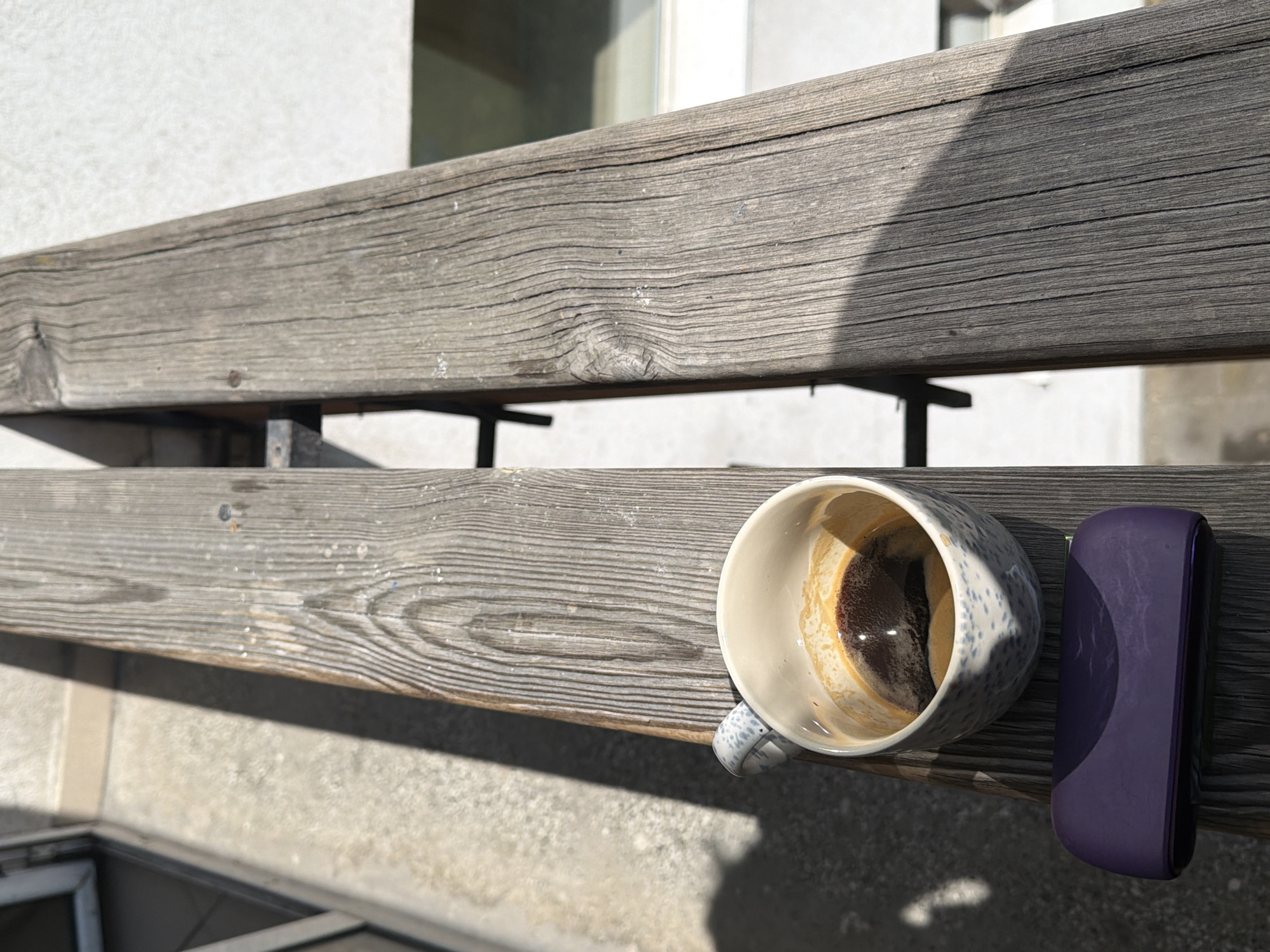 The image shows a partially consumed cup of coffee and a small, dark purple, rectangular object resting on a weathered, grey wooden balcony railing. The railing consists of two horizontal planks supported by dark metal brackets. The background includes a textured white wall and glimpses of a window. The location is likely Sofia, Bulgaria, based on architectural style and common urban features. No people are visible. The scene suggests a casual, relaxed setting, possibly a residential balcony.