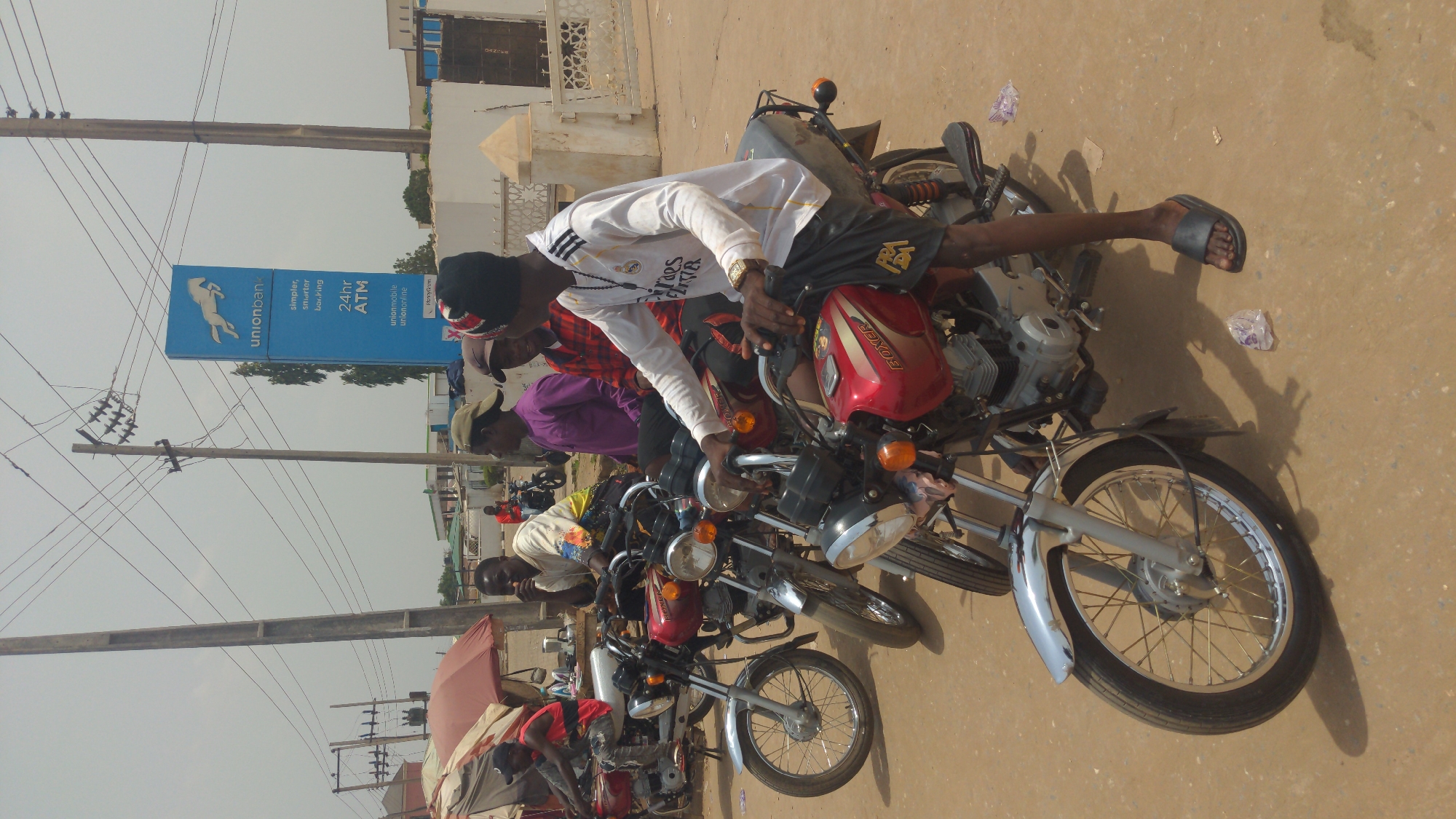 The image depicts an outdoor scene, likely a busy street or commercial area in Ado, Nigeria, during daylight hours. The ground is unpaved and appears dusty, with scattered pieces of litter. The bright sunlight casts distinct shadows, indicating a clear, sunny day.
The primary subjects are several men gathered around multiple motorcycles. In the foreground, a young man wearing a white long-sleeved shirt with Adidas stripes and "Fly Emirates" branding, dark shorts, and sandals, is seated on a red motorcycle. He also wears a dark cap and a wristwatch. Another man in a red and black plaid shirt and a third in a purple shirt are visible behind him, also near motorcycles. Further back, more men and motorcycles can be seen, some partially obscured. The motorcycles appear to be commuter or commercial models, with one prominent red motorcycle clearly displaying "BOXER" on its fuel tank.
In the background, a tall, blue vertical sign for "Unionbank" stands prominently. The sign features a horse logo and text that reads "unionbank," "simpler smarter banking," "24hr ATM," and information about "unionmobile" and "uniononline," along with a "Hotline." Utility poles with numerous overhead wires crisscross the sky, suggesting an urban or semi-urban setting. Buildings with light-colored facades are visible behind the bank sign and other structures. The overall impression is one of a casual gathering point, possibly a motorcycle taxi stand or a common waiting area on a street.