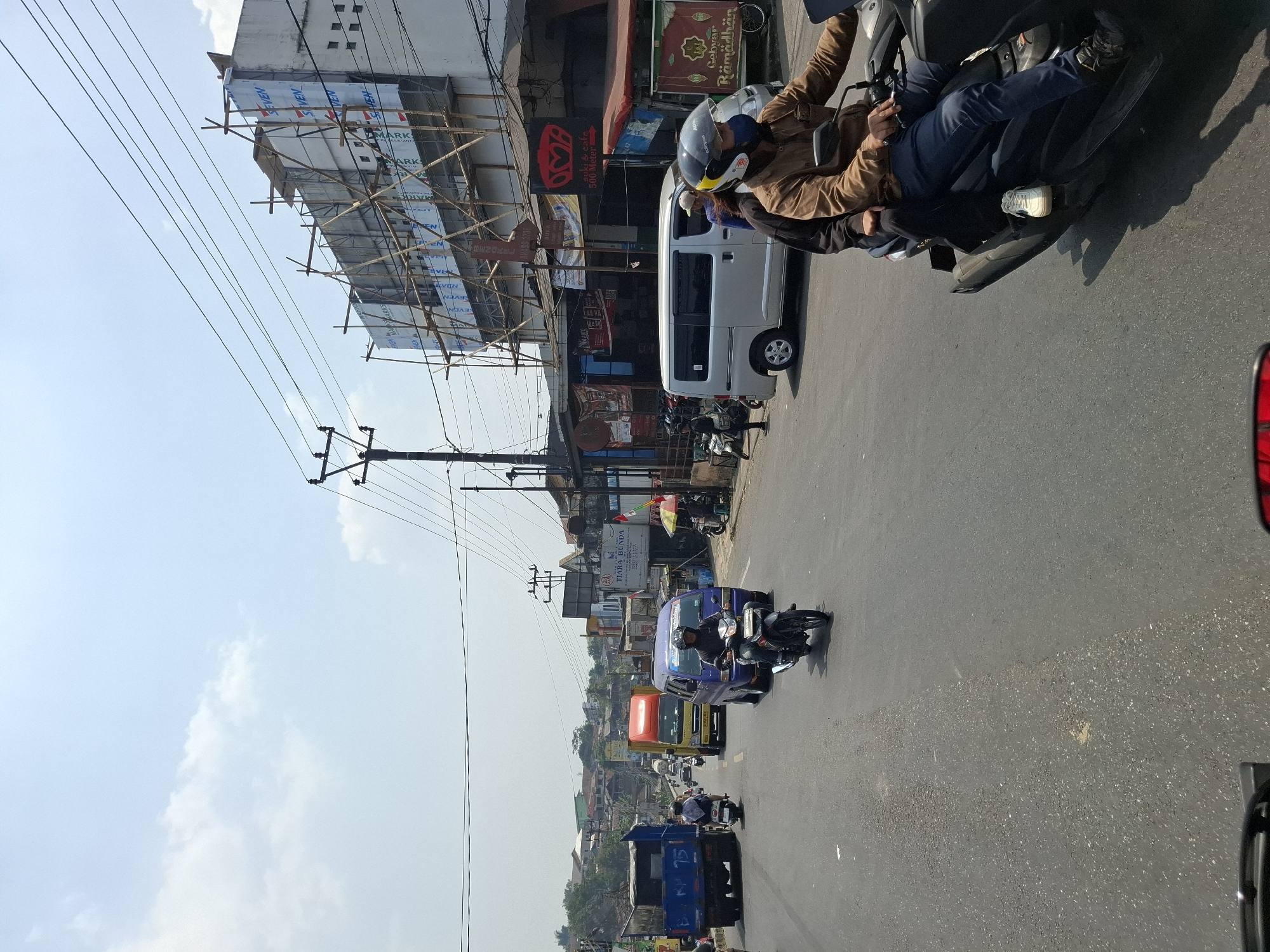 A daytime exterior view shows a street scene in Cibadak, Indonesia, with moderate vehicular traffic. In the foreground, a dark-colored scooter carries two individuals. The rider, wearing a brown jacket, blue jeans, and a dark helmet, operates the scooter. The passenger, dressed in dark clothing, sits behind the rider. The scooter is positioned on the right side of the paved road, which has visible lane markings and some wear and tear.
On the road, several vehicles are present. To the left, a blue truck with the number "75" visible on its side is driving away from the camera. Ahead of it, a yellow and orange truck is also in motion. A purple van is traveling in the same direction as the scooter. Several motorcycles are also on the road, with one rider in the foreground on a black motorcycle wearing a dark helmet and blue shirt, and another rider further back on a white motorcycle.
The environment includes buildings lining the right side of the road. One building is undergoing renovation or construction, covered in scaffolding and white sheeting with "SEVEN" printed on it. Other buildings appear to be commercial establishments, with signs that are partially visible. A street pole with electrical wires runs overhead. The sky is clear with scattered white clouds, indicating a sunny day. The overall impression is a busy urban street environment.