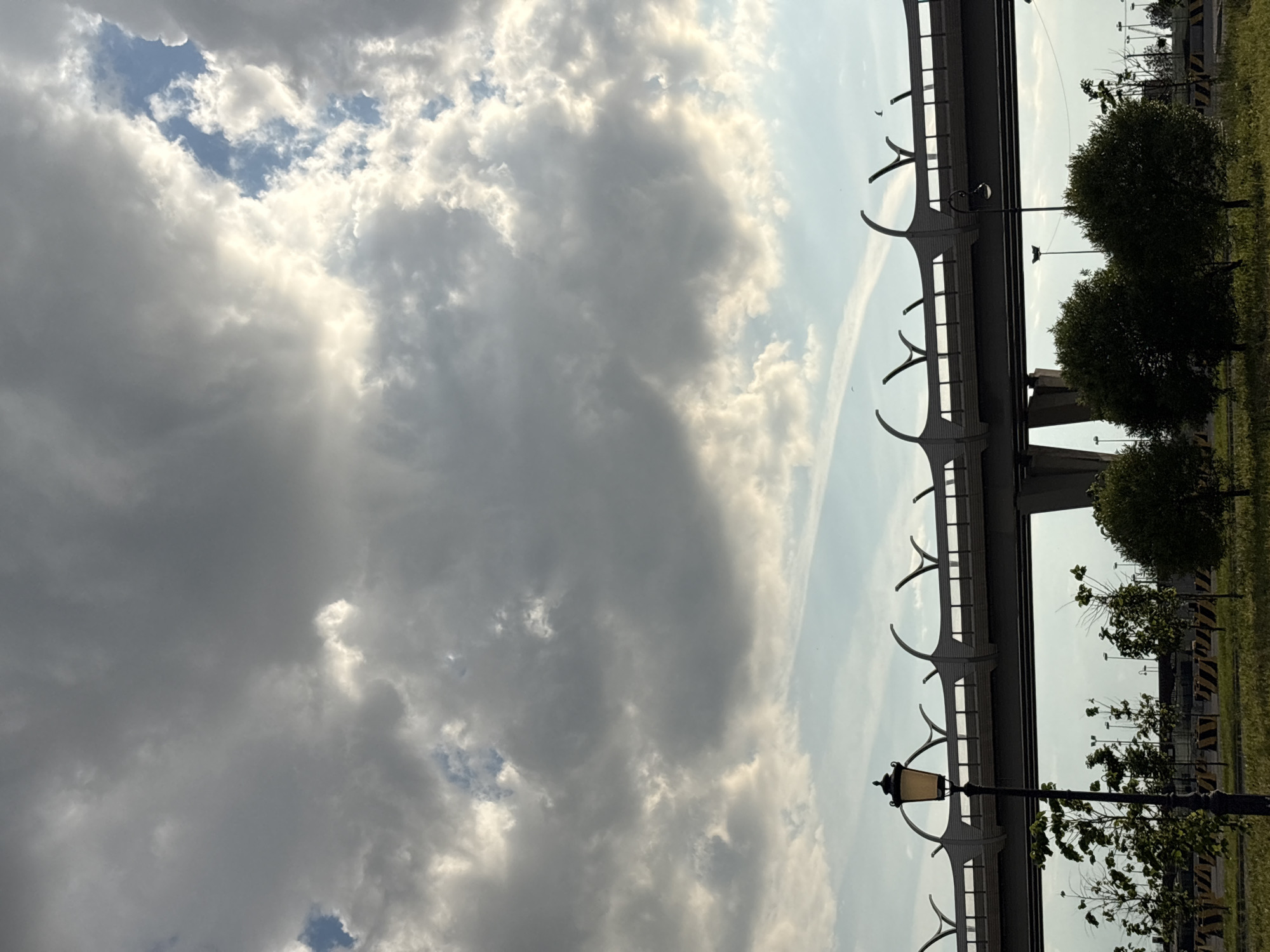 The image, oriented sideways, presents a dynamic sky above a prominent overpass structure. The upper left portion of the frame is dominated by a cloud-filled sky. Large, textured clouds vary in color from bright white where sunlight breaks through to darker, heavy grey masses. Several patches of clear blue sky are visible between the cloud formations. A faint white contrail is also discernable in the lighter sky area on the right.
A substantial concrete bridge or overpass runs horizontally across the middle of the image. Its underside is characterized by a repeating series of decorative, white, inverted arch-like elements that create a distinctive pattern against the sky. Below the bridge and extending towards the right, urban landscaping is visible. A classic-style black street lamp with an ornate, yellowish glass lantern stands in the lower right foreground. Green bushes and trees with dense foliage are scattered beneath and around the bridge structure, alongside grassy areas. Further in the background, beyond the main bridge, a road or paved area is faintly visible with yellow and black striped safety barriers.
The scene takes place during the day, likely late afternoon, indicated by the strong highlights on the clouds from the sun. The weather is partly cloudy. No human activity or visible text is present in the image. The infrastructure depicted is typical of a modern urban environment, consistent with locations such as Saint Petersburg, Russia.