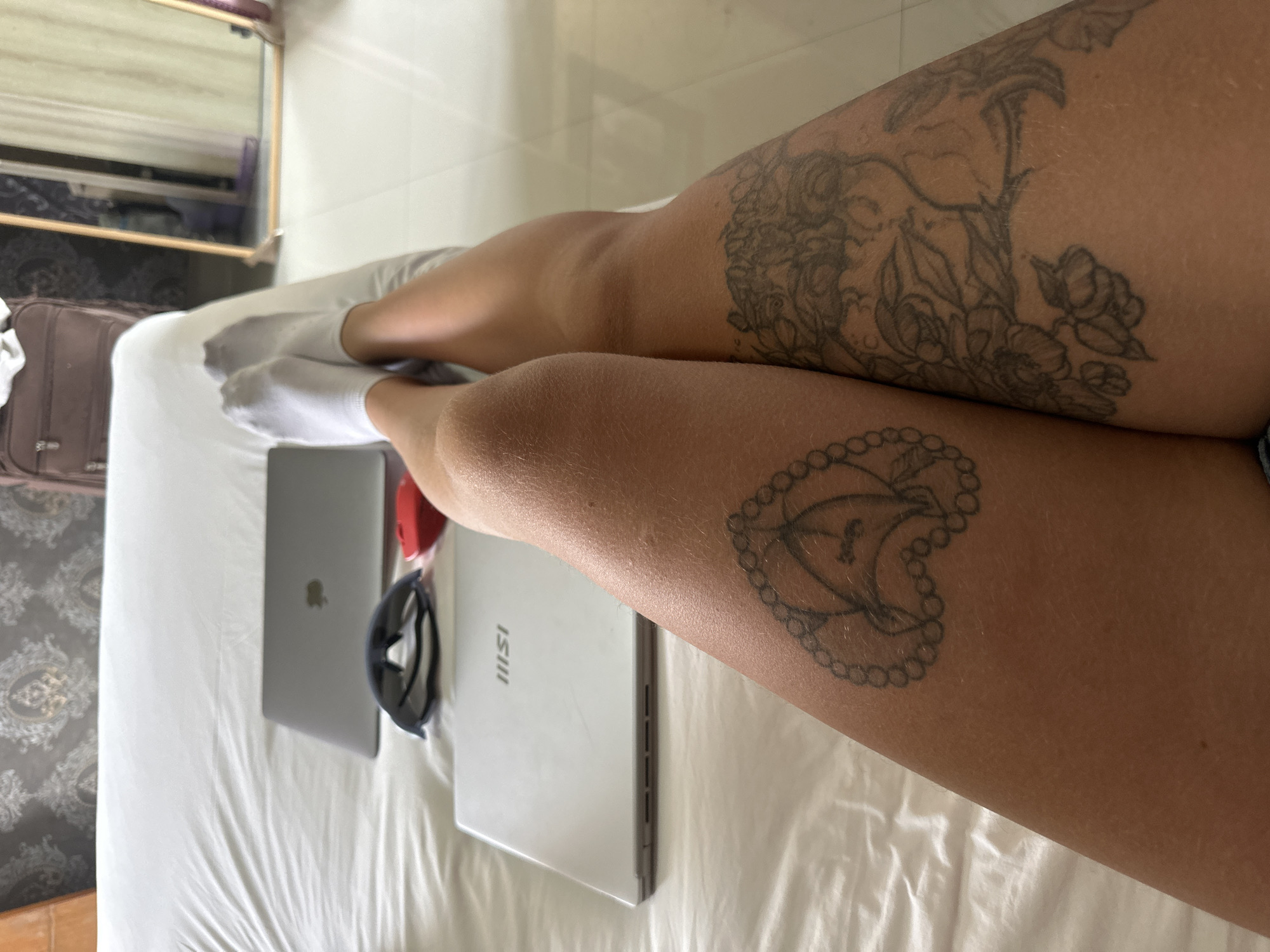 In an indoor setting, likely a bedroom, a person is lying on a white bed with their legs crossed and extended towards the camera. The person is wearing white socks. There are two laptops on the bed, one silver MSI laptop and one silver MacBook. Next to the MacBook are a pair of dark sunglasses and a red computer mouse. The person has tattoos on their thighs. The left thigh has a tattoo of a heart outline with a detailed design inside, and the right thigh has a floral tattoo. The background shows a patterned wallpaper and a suitcase, with a mirror and a doorway visible in the distance. The flooring appears to be white tiles.