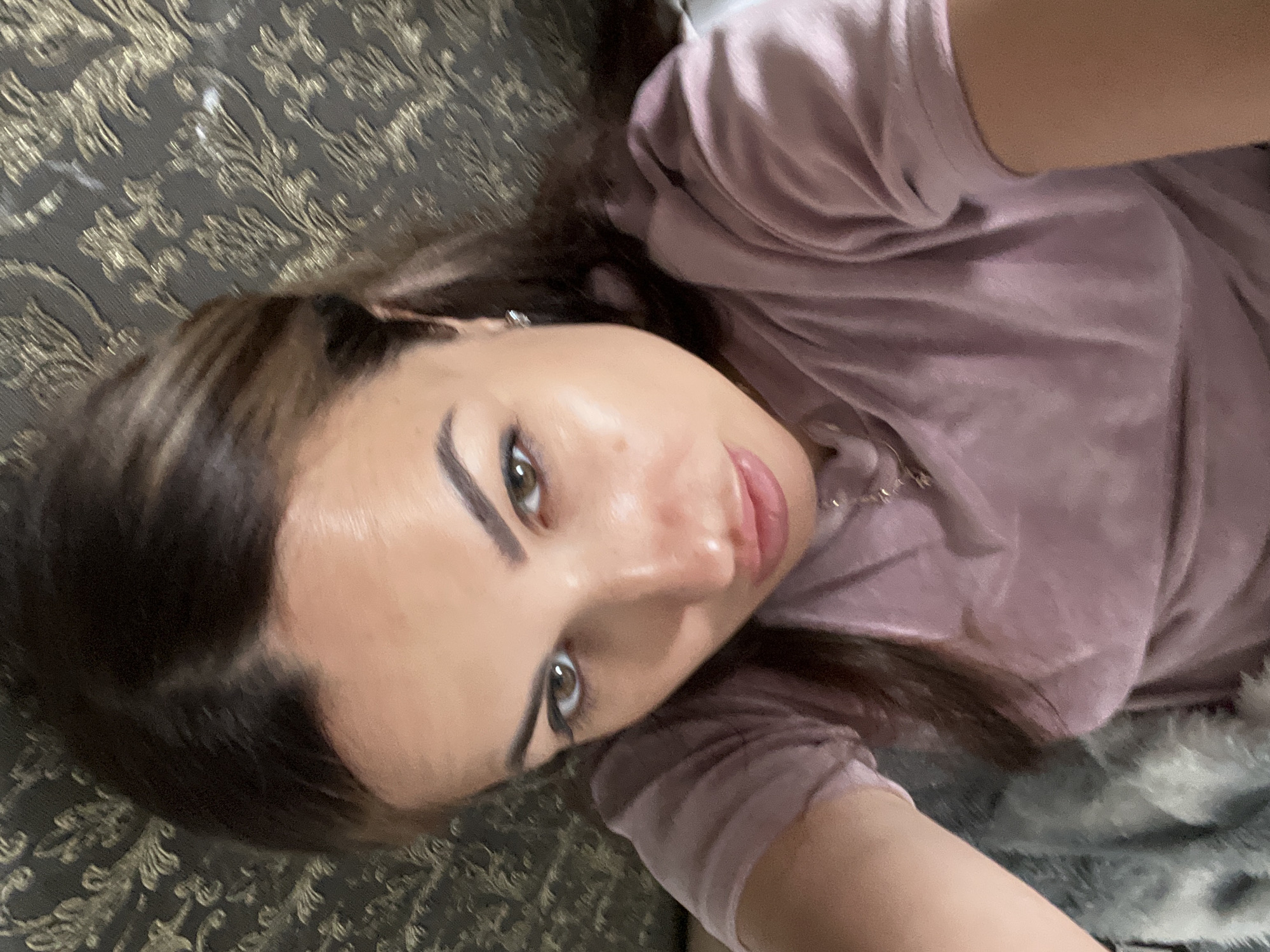 A woman with brown hair and brown eyes is looking at the camera. She is wearing a pink t-shirt. The background is a patterned fabric.