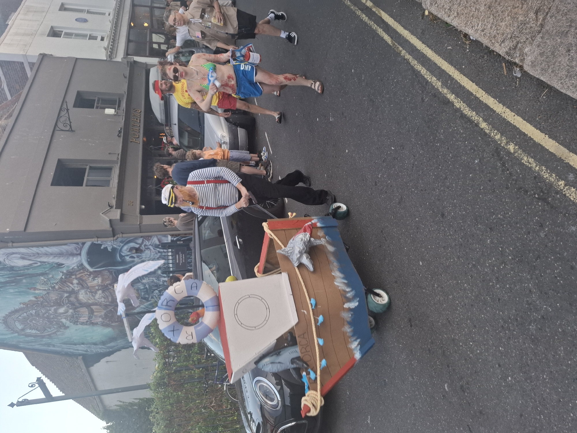 In St Leonards, United Kingdom, on a street with double yellow lines and a grey curb, a man in a striped shirt, red suspenders, and a white sailor hat with a pipe in his mouth pushes a small boat on wheels. The boat is decorated with blue and white waves, a life preserver that reads "ORCA," and several small blue fish. The boat is made of cardboard and has a white box structure inside. Behind him, a woman in a bikini and blue shorts, with what appears to be fake blood splattered on her skin, walks while holding a water gun. To her right, a man in a tan shirt and shorts holds a bottle. In the background, buildings line the street, including one with a mural on its side and another with "PORTERS" signage above its entrance. Other people are visible in the background, and a black car is parked to the left of the man with the boat.