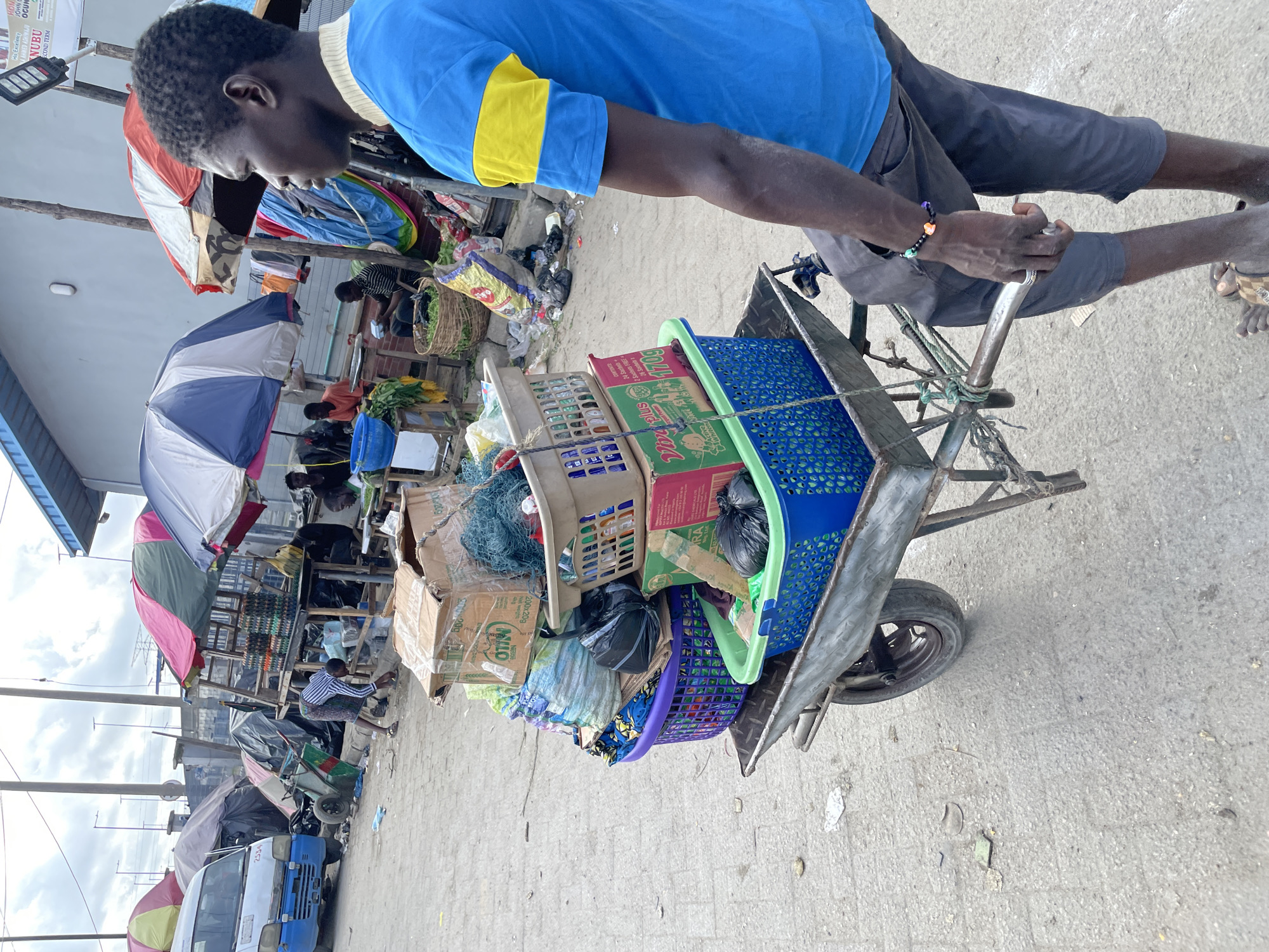 In the city of Lekki, Nigeria, a young man is seen pushing a wheelbarrow loaded with various goods. The wheelbarrow contains several baskets and boxes, one of which is a brown cardboard box labeled "Milo". Other items include a green net, a purple basket filled with fabric, and a blue basket holding more goods. The man is wearing a blue t-shirt with yellow stripes on the sleeves and dark grey trousers. He has a bracelet on his left wrist and a ring on his left hand. The background reveals a bustling market scene with other people, stalls with umbrellas, a parked van, and various scattered debris on the ground. The overall environment suggests a busy outdoor marketplace under a cloudy sky.