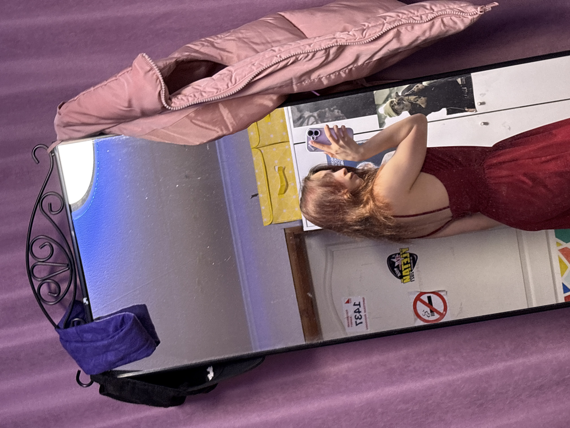 A woman in a red dress takes a selfie in a mirror. The mirror is on a purple blanket. A pink puffy jacket is draped over the top corner of the mirror. A blue bag hangs off the left side of the mirror. In the reflection, a dresser is visible as well as a door with stickers. The stickers include one that says “NATEIV” and one that says “NO SMOKING.” A sticker that says “T43J” is also visible. The location is Прикамский, Russia, based on the "T43J" sticker, which is a regional code.