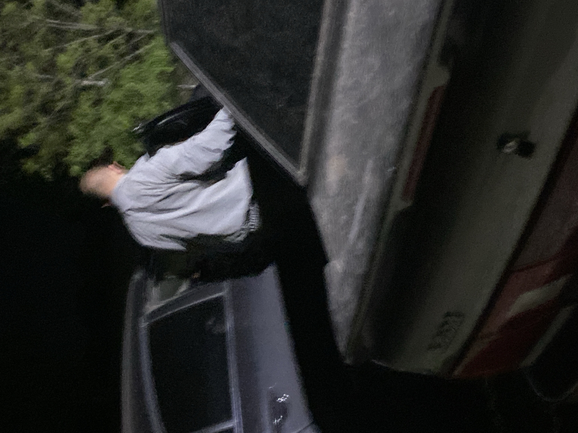 The image shows a person with a shaved head, wearing a light-colored shirt and dark pants, possibly leaning into the open driver's side of a truck or similar vehicle. The vehicle is partly visible with dark and reddish-brown sections, potentially a dent on the right side. Trees and darkness suggest an outdoor, possibly wooded environment. The orientation of the image is not clear, so determining if the vehicle is damaged or overturned is difficult. There is no information in the image to suggest a location in Ustyvytsia, Ukraine.