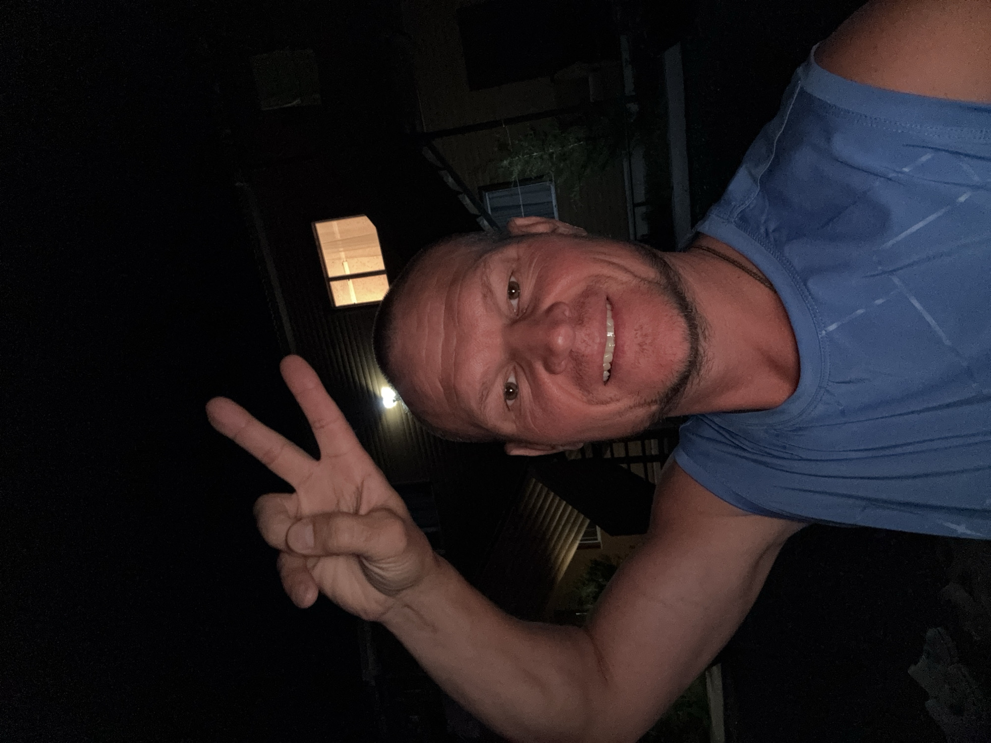 A man in a blue sleeveless shirt poses for a selfie at night, holding up two fingers in a peace sign. He has a shaved head, a clean-shaven face with a slight stubble on his chin, and is smiling. In the background, a building with lit windows is visible. The image appears to have been taken outdoors. The location is identified as Zharbulak, Kazakhstan.