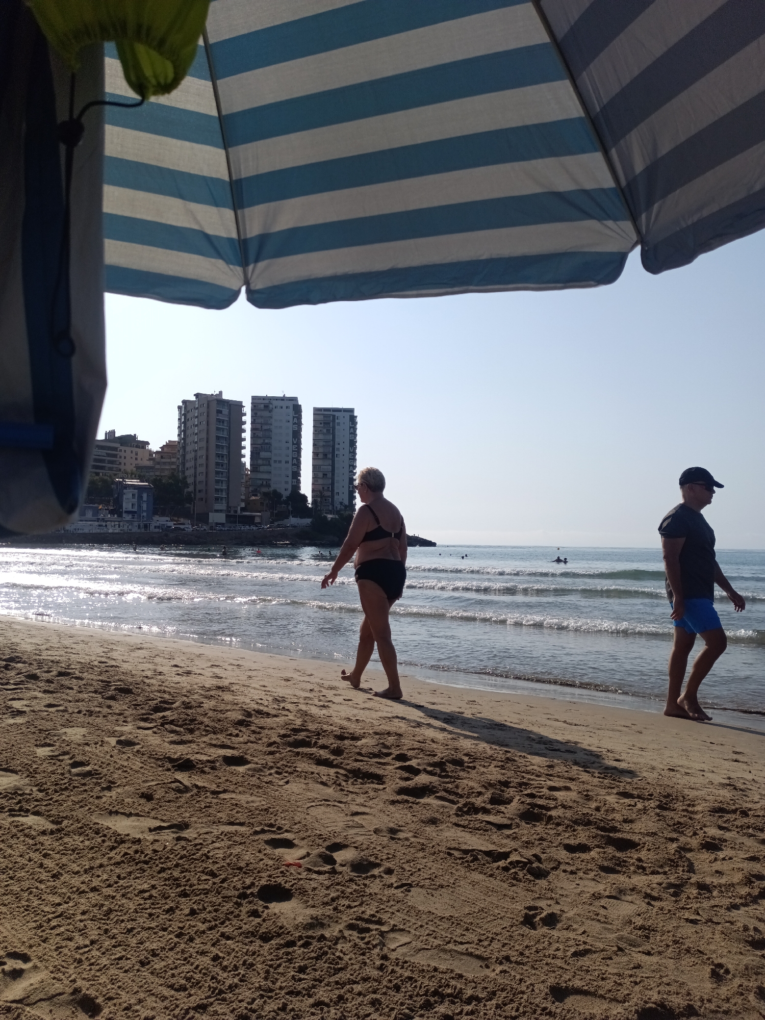 An eye-level, wide shot captures a beach scene in Orpesa, Spain, under a striped blue and white beach umbrella. In the foreground, tan sand is covered with numerous footprints. Two people are visible walking on the sand. To the left, a woman in a black bikini walks towards the right, her body angled away from the camera. To the right, a man wearing a dark t-shirt and blue swim trunks walks towards the right, his body angled away from the camera and his face obscured by a dark baseball cap. In the background, the ocean is calm with gentle waves and sparkling sunlight. Several people are visible in the water and in the distance. Along the coastline, a row of multi-story buildings, including hotels and apartments, are situated. The sky is clear and bright.
