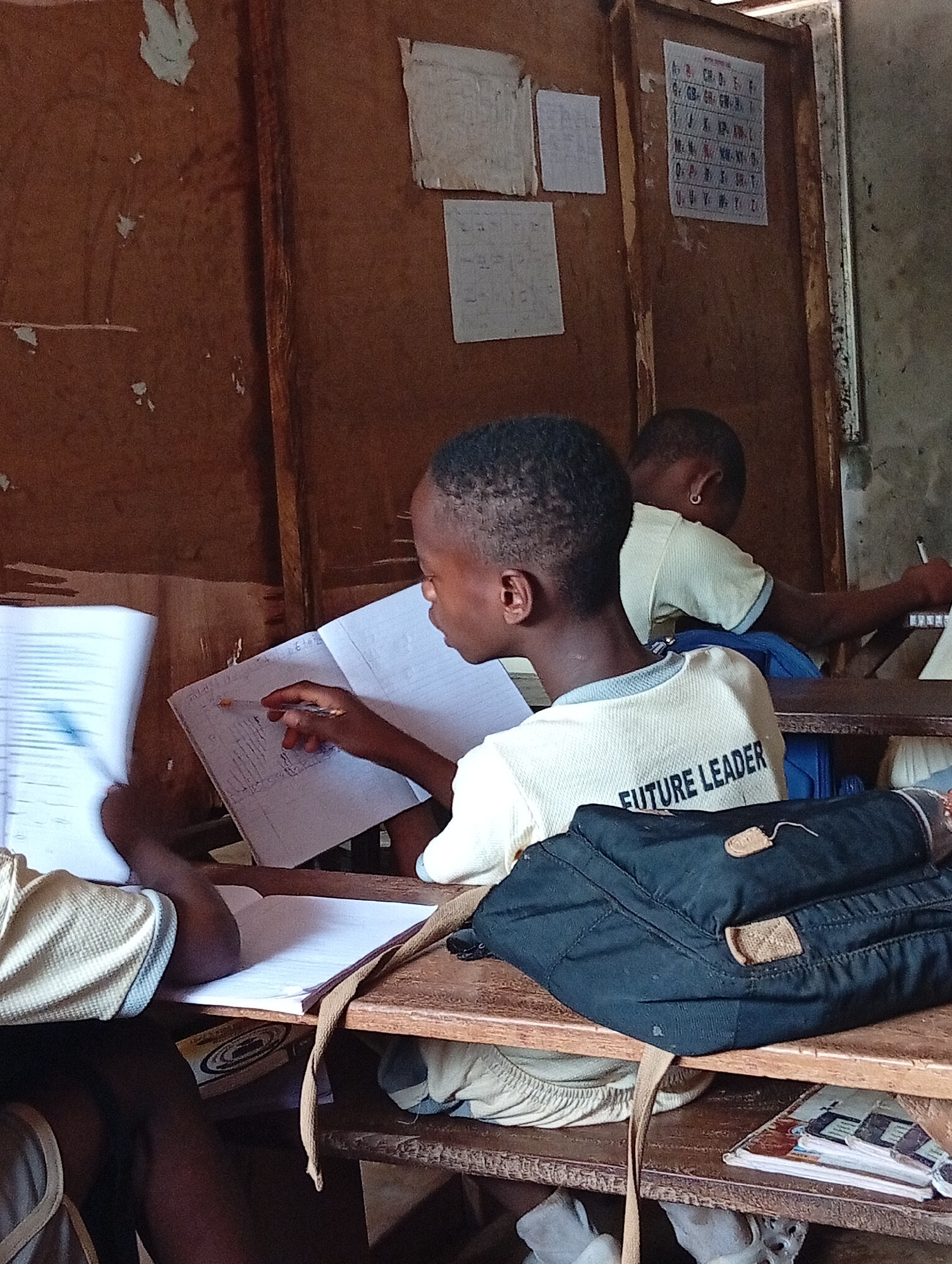 In Ukwu-Abwa, Nigeria, a classroom setting features three young students. One, in the foreground, wears a shirt with "FUTURE LEADER" printed on the back. He is seated at a wooden desk, intently working on a notebook. Another student, partially visible behind him, is also writing. A black bag rests on the desk in front of the "FUTURE LEADER" student. Behind them, the classroom wall is adorned with papers and charts.