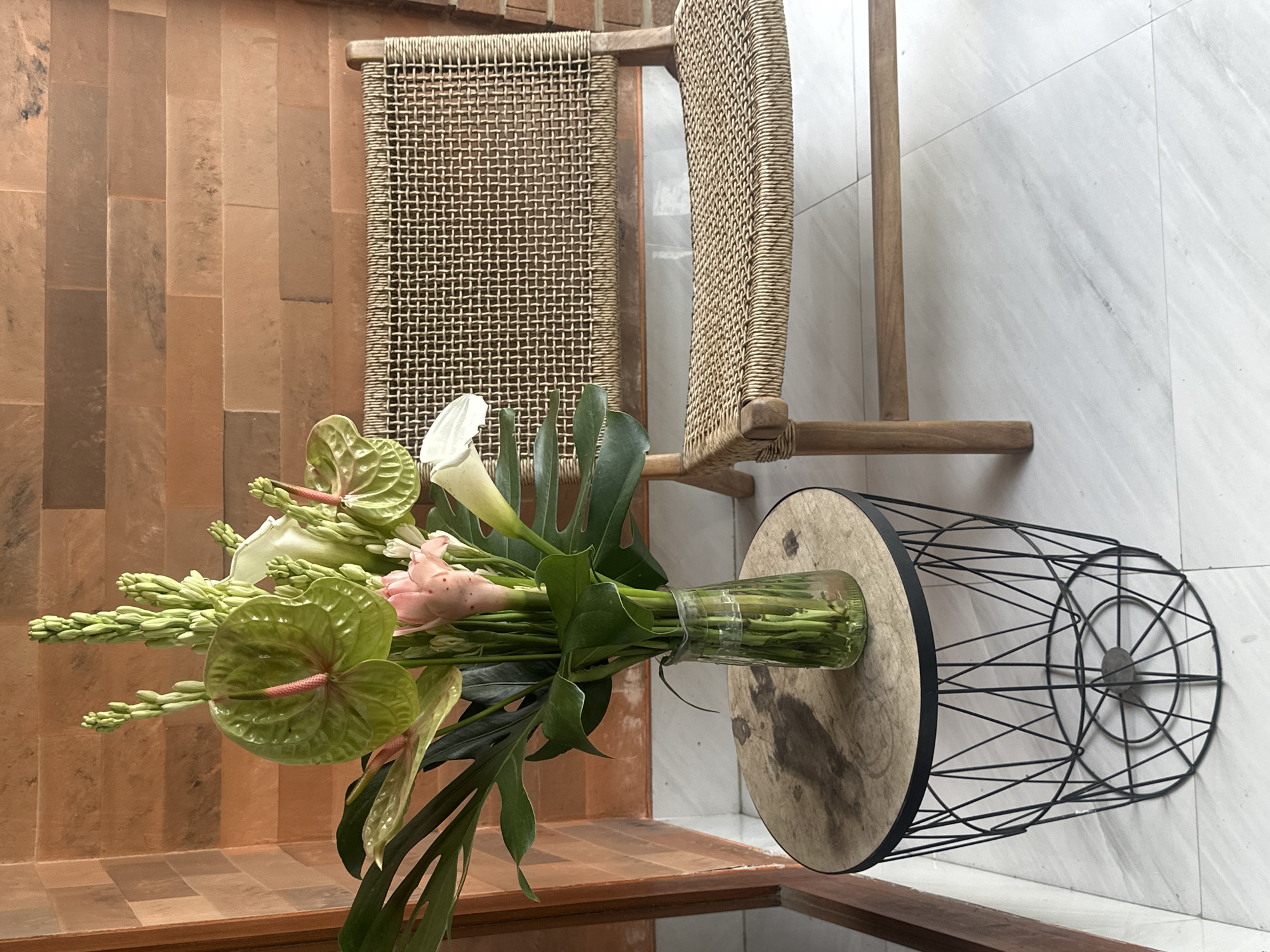 Here is a description of the image:
The image shows a still life scene featuring a floral arrangement, chairs, and a side table.
The floral arrangement includes green anthuriums, calla lilies, and other flowers, arranged in a glass vase on a round wooden side table with a black metal cage base. The table is placed in front of two woven chairs, one with a more open weave and the other with a tighter weave. The background consists of two walls: one tiled in a vertical, reddish-brown pattern and the other in a light-colored marble or tile. The scene is located in Mengwi, Indonesia.
