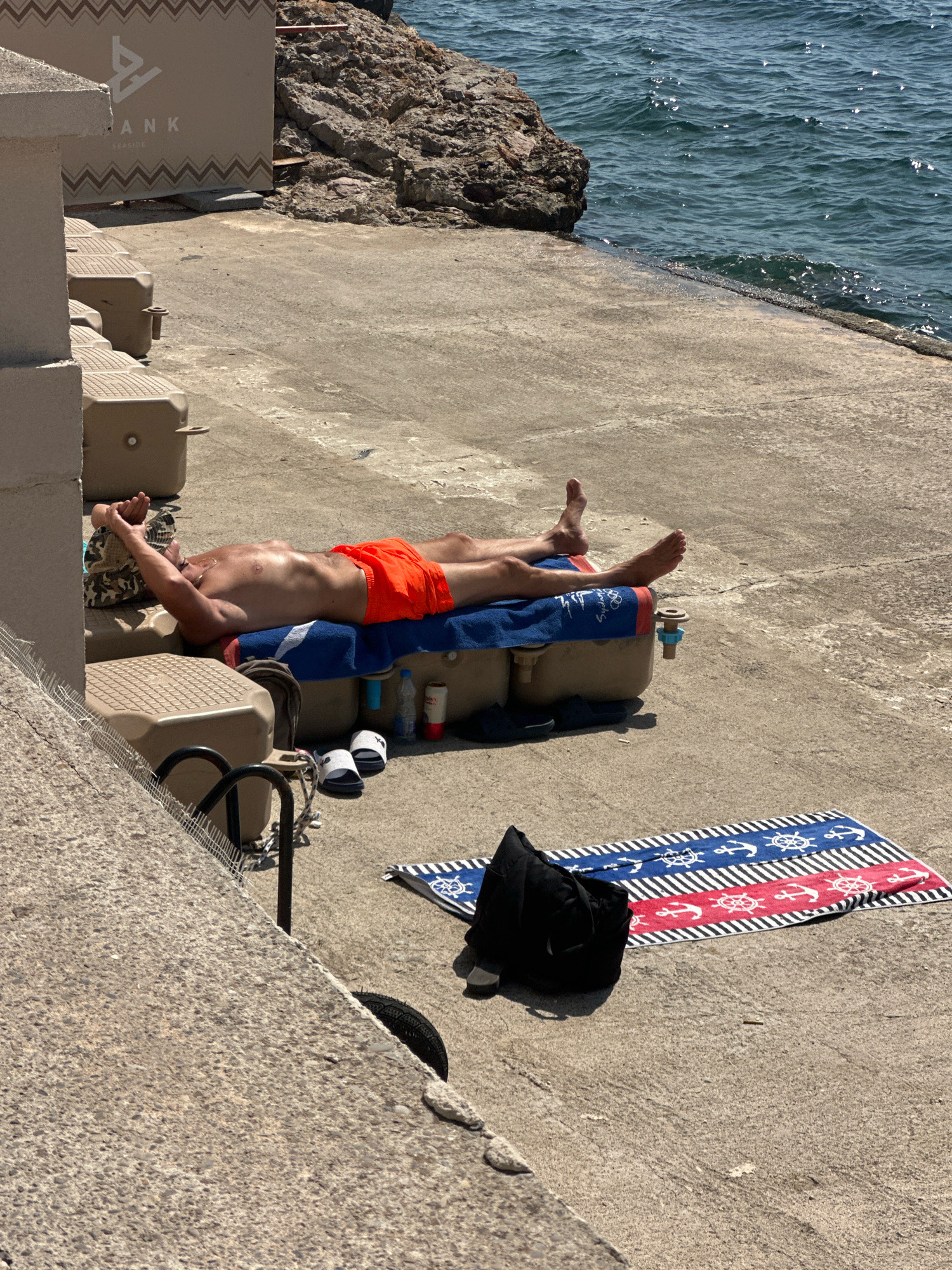 A man is sunbathing on a concrete pier next to the sea in Budva, Montenegro. He is lying on his back, wearing orange swim trunks and a camouflage hat. He has his arms above his head, with his hands behind the hat. A blue towel is spread out on a large, tan-colored floating platform beneath him. To his right are a bottle of water and a white and red can. Nearby on the concrete are a pair of white flip-flops, a black bag, and a striped towel with anchor motifs. In the background, the turquoise sea is visible with small waves, and a rocky outcropping. Behind the floating platform is a tan-colored structure with the word "BANK" visible.