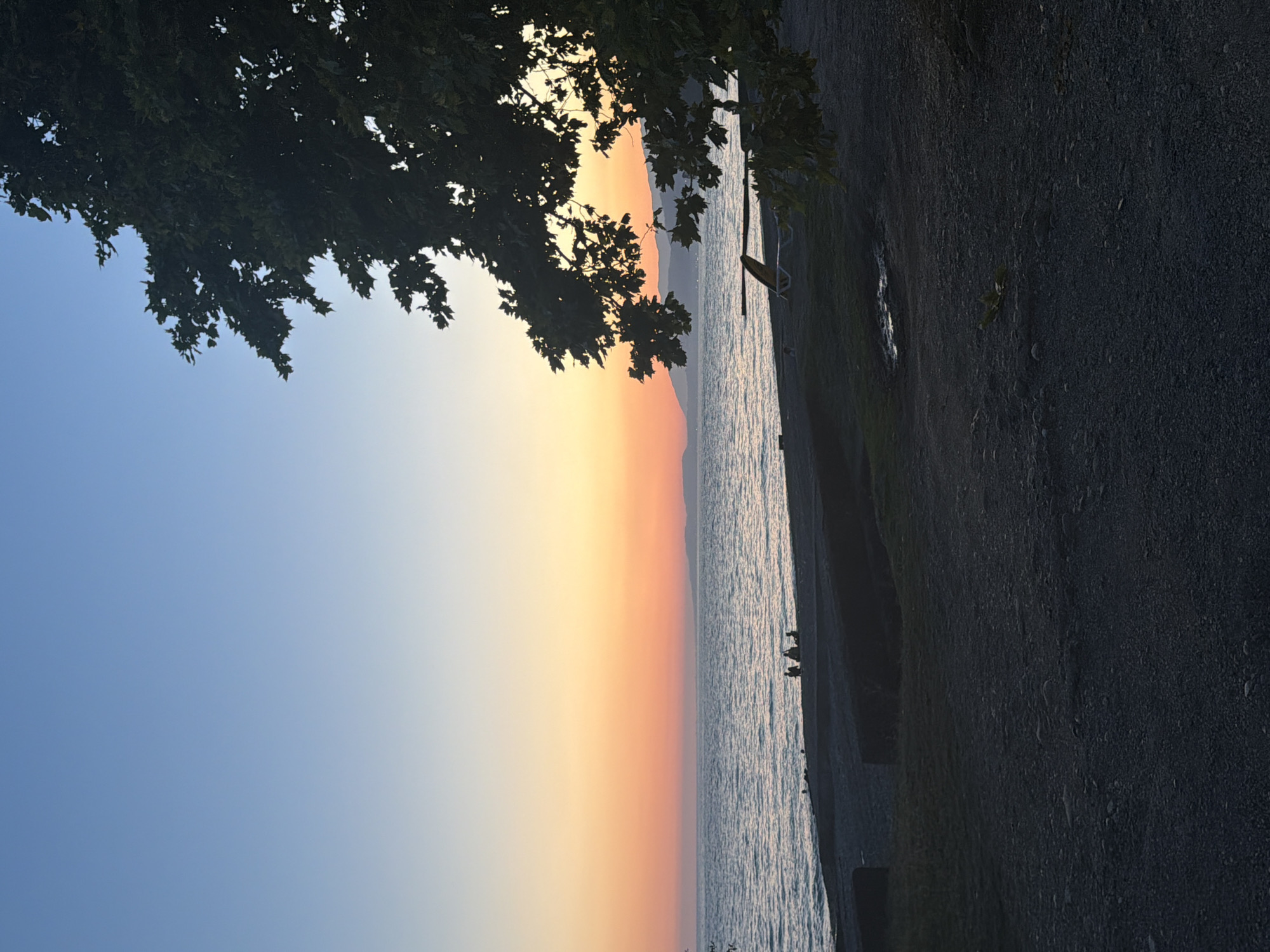 A serene sunset scene unfolds at a beach in Eshyra, Abkhazia. The sky transitions from a clear, soft blue at the top to a gradient of orange and pink hues near the horizon, creating a warm and tranquil atmosphere. The sun is not directly visible, but its light illuminates the sky and reflects on the calm, rippling water of the sea.
In the foreground, a dark, pebbly beach stretches out, with patches of grass and some scattered rocks. A few dark, indistinct shapes, likely people, are gathered near the water's edge, appearing to be sitting or standing in conversation. Further back on the beach, a lone lounge chair is visible, suggesting a place for relaxation.
A large, leafy tree with dark green foliage dominates the right side of the frame, its branches silhouetted against the colorful sky. The tree's leaves are partially illuminated by the setting sun. In the distance, a hazy line of mountains or hills can be seen against the horizon, adding depth to the landscape. The overall impression is one of peaceful evening by the sea.