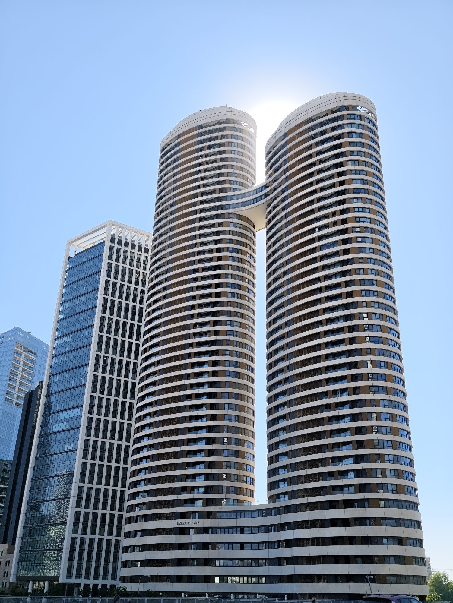 This is a low-angle, full-frame, eye-level shot of the futuristic residential towers known as "The Twins" in Tel Aviv, Israel, with a clear blue sky and the sun prominently positioned behind the upper portion of the left tower, creating a lens flare effect. The buildings are characterized by their modern, curvilinear architecture, with the two primary towers connected by a skybridge.
The left tower, partially obscured by the sun, features a white exterior with horizontal banding of dark windows and balconies. The right tower is a mirror image of the left, with a similar white exterior and dark window bands. The lower section of the towers exhibits a more curved, undulating design, with numerous horizontal bands of windows.
To the left of "The Twins," a tall, rectangular skyscraper with a white grid-like facade and large, dark glass panels is visible. Further to the left and slightly behind, another glass skyscraper with a blue tint and white horizontal accents can be seen. The very bottom of the frame shows the top of a barrier or railing, with faint hints of trees and a car visible beyond. The overall scene depicts a modern urban landscape with an emphasis on architectural innovation.