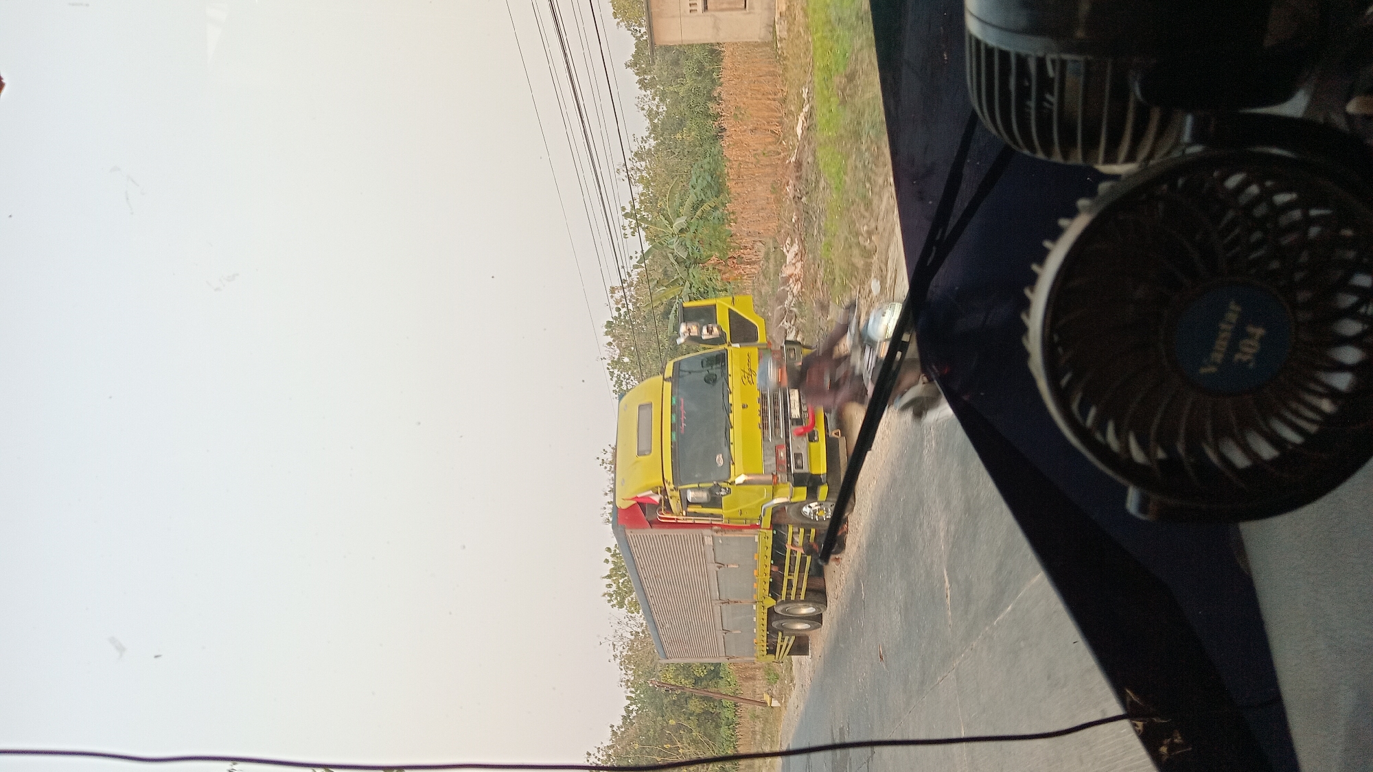 This is a photograph taken from inside a vehicle, likely a bus or truck, looking out the windshield. The perspective shows the dashboard in the foreground, with a small black fan with the text "Vanstar 304" visible on it. The windshield has a reflection that obscures some of the view.
Outside the vehicle, on a paved road, a bright yellow truck with a silver cargo box is parked. The truck has an open driver's side door and a red flag mounted on its roof. A person on a motorcycle, wearing a helmet, is riding past the truck on the road. The motorcycle rider is blurred due to motion.
The environment appears to be rural or semi-rural, with dry vegetation, trees, and a small building visible in the background. Power lines stretch across the sky. The time of day seems to be late afternoon or early morning due to the soft, hazy lighting.
The provided information about the location being "Klokah, Indonesia" is noted, but the image itself does not contain explicit visual cues that pinpoint this specific location. However, the scene depicts a typical road in a warm climate, consistent with Southeast Asia.