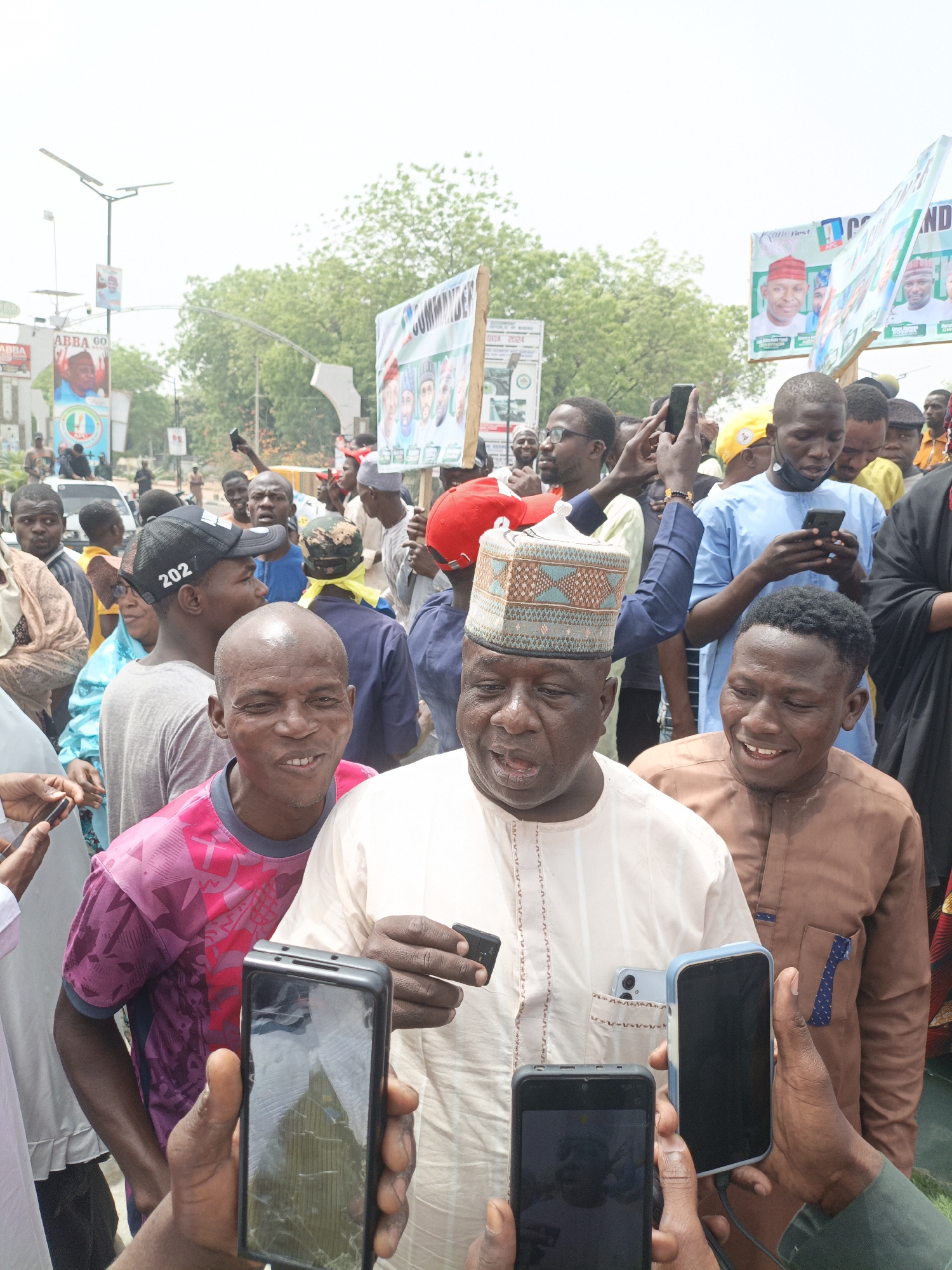 The image captures a scene in what appears to be a public gathering, possibly a political event or rally, in Giginyu, Nigeria. A middle-aged man in a traditional patterned hat and light-colored robe is the focal point, surrounded by a crowd of people. Many individuals are holding up smartphones, presumably to record or photograph the central figure. The man seems to be speaking or gesturing. The crowd is diverse in terms of age and attire.
Background elements include banners and signs with images of political figures, indicating the political nature of the event. The environment appears to be an urban setting with streetlights, buildings, and trees visible. The image is well-lit, suggesting it was taken during daylight hours.