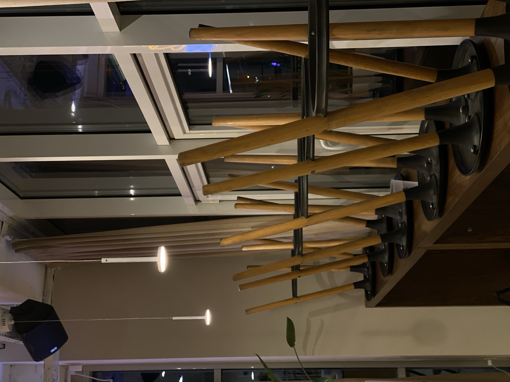 The image shows a row of bar stools stacked upside down on top of a wooden counter, likely in a closed establishment. The stools have wooden legs and black seats. In the background, large windows reflect the interior and exterior lights. There is a black speaker mounted on the ceiling and two modern pendant lights hanging down. A large potted plant with green leaves is visible to the left. The environment appears to be indoors, possibly a cafe or bar, with ambient lighting and a muted color palette. The scene is devoid of people. The image does not provide enough information to determine its specific location in Vishnyaki, Russia.