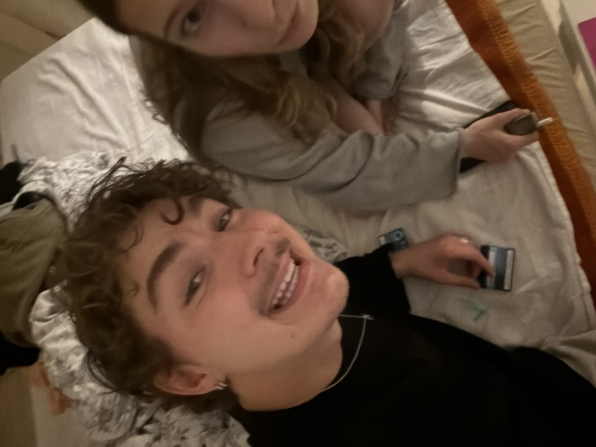 The image shows two young adults lying on a bed. The person on the left, with curly brown hair and a mustache, is looking towards the camera and smiling. They are wearing a black t-shirt and a silver necklace. A few small objects are visible on the bed near them, including what appears to be a credit card. The person on the right has long brown hair and is wearing a grey sweatshirt. They are partially visible and their face is turned away from the camera. The environment appears to be a bedroom, with white bedding and some patterned fabric visible in the background. There is no information in the image that would indicate the location as Vracar Urban Municipality, Serbia.