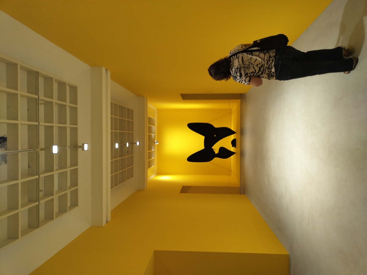 The image depicts a person walking down a brightly lit, yellow-walled corridor towards a large black silhouette of a dog's head with a small mouse visible within its negative space. The corridor has a white, grid-like ceiling with recessed lighting. The floor is a smooth, light gray concrete. The person, a woman with dark hair, is wearing a patterned t-shirt and dark jeans, and is carrying a black bag over her shoulder. She is walking away from the camera, looking towards the artwork. This artwork is likely part of an exhibition at an art gallery or museum. Given the distinctive yellow walls and minimalist aesthetic, this could be the Tel Aviv Museum of Art, which is known for its modern architecture and bright interior spaces, and has hosted numerous contemporary art exhibitions.