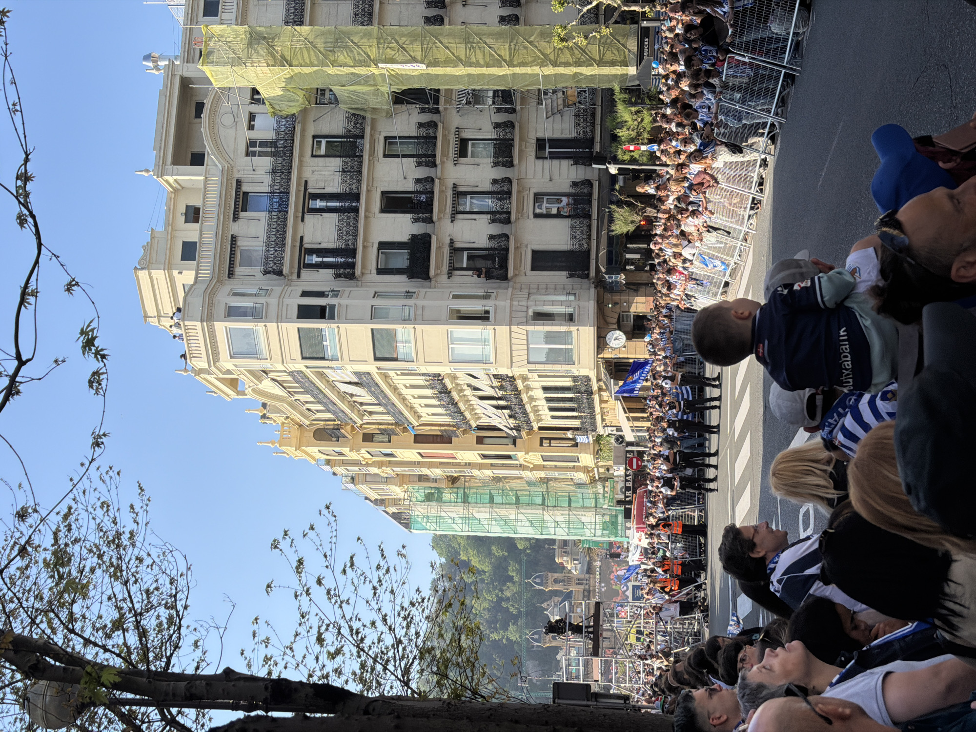 The image captures a vibrant daytime scene in a city street, likely San Sebastián, Spain. A large crowd of spectators lines the street behind metal barriers, observing an event or parade. Many individuals in the crowd, including a child on an adult's shoulders in the foreground, are wearing blue and white striped shirts, suggesting a sports-related celebration, possibly for a local team like Real Sociedad. The child's shirt has "Xabaliak" partially visible on the back.
The street is flanked by grand, multi-story buildings with ornate balconies and architectural details. One prominent building on the left features extensive scaffolding covered in light green netting, indicating ongoing construction or renovation. The clear blue sky suggests pleasant weather. In the background, green hills are visible, adding a natural element to the urban landscape.
Further down the street, a clock is visible on one building, and a small "STARBUCKS" sign can be partially seen. An awning on a building displays "EL BAR BAS." People are also observed on some of the balconies, enjoying the view of the event below. In the mid-ground, a stage or platform with equipment is set up, further indicating an organized public gathering. Trees with sparse, budding leaves frame the left side of the shot, contrasting with the man-made structures. The overall atmosphere appears festive and engaging.