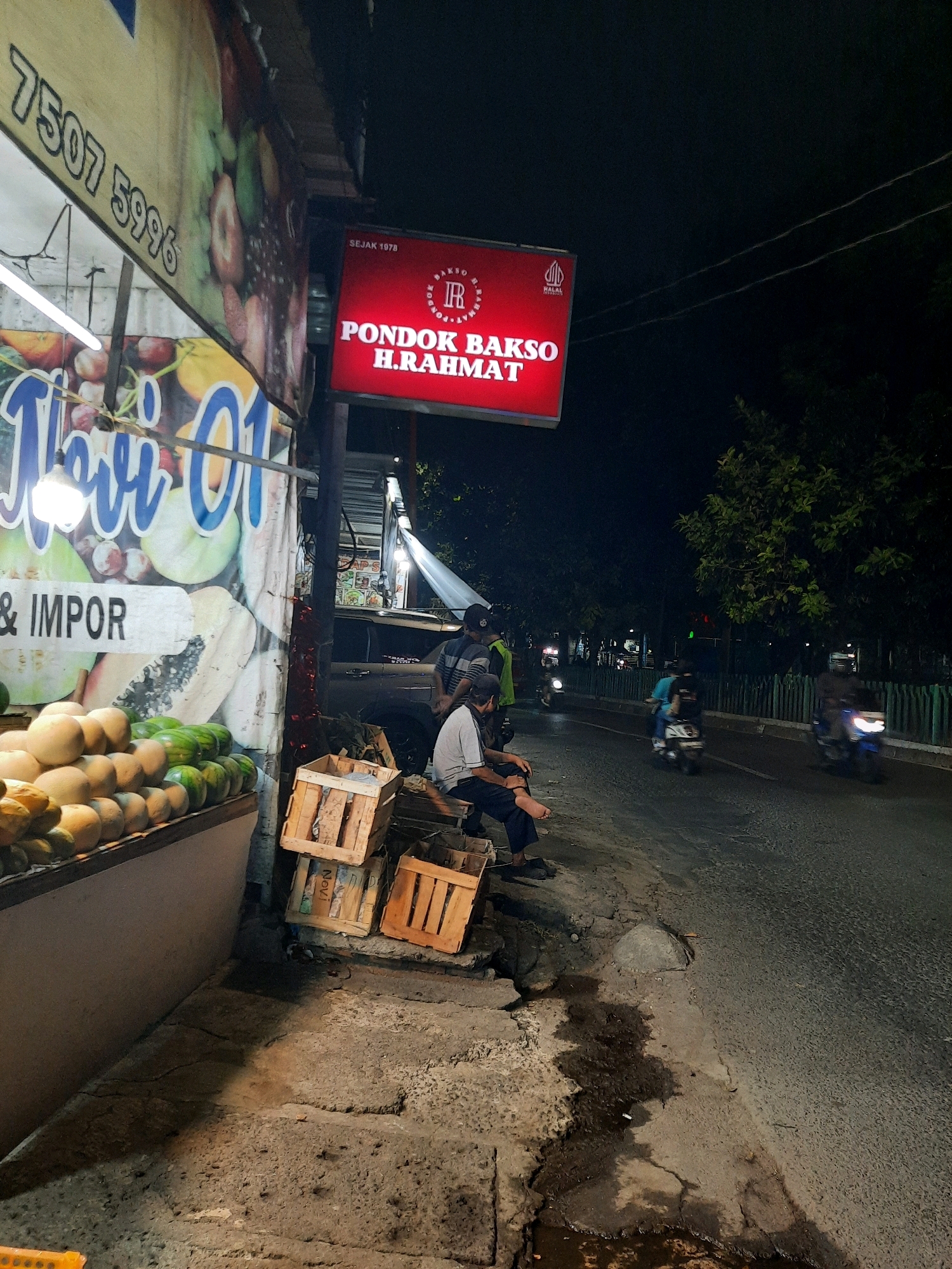 The image depicts a street scene at night in East Jakarta, Indonesia. On the left side of the frame, a fruit stand displays rows of melons and watermelons. A banner with the text "7507 5996" and "TOS 01 & IMPOR" is visible above the fruits. A red illuminated sign for "PONDOK BAKSO H.RAHMAT" with the text "SEJAK 1978" is prominently displayed. In front of the fruit stand, several wooden crates are stacked. A man is seated on the crates, facing away from the camera. Further down the street, a busy road with several motorbikes is visible. Trees and a green fence line the side of the road. The overall impression is of a lively street scene with commercial activity at night.
