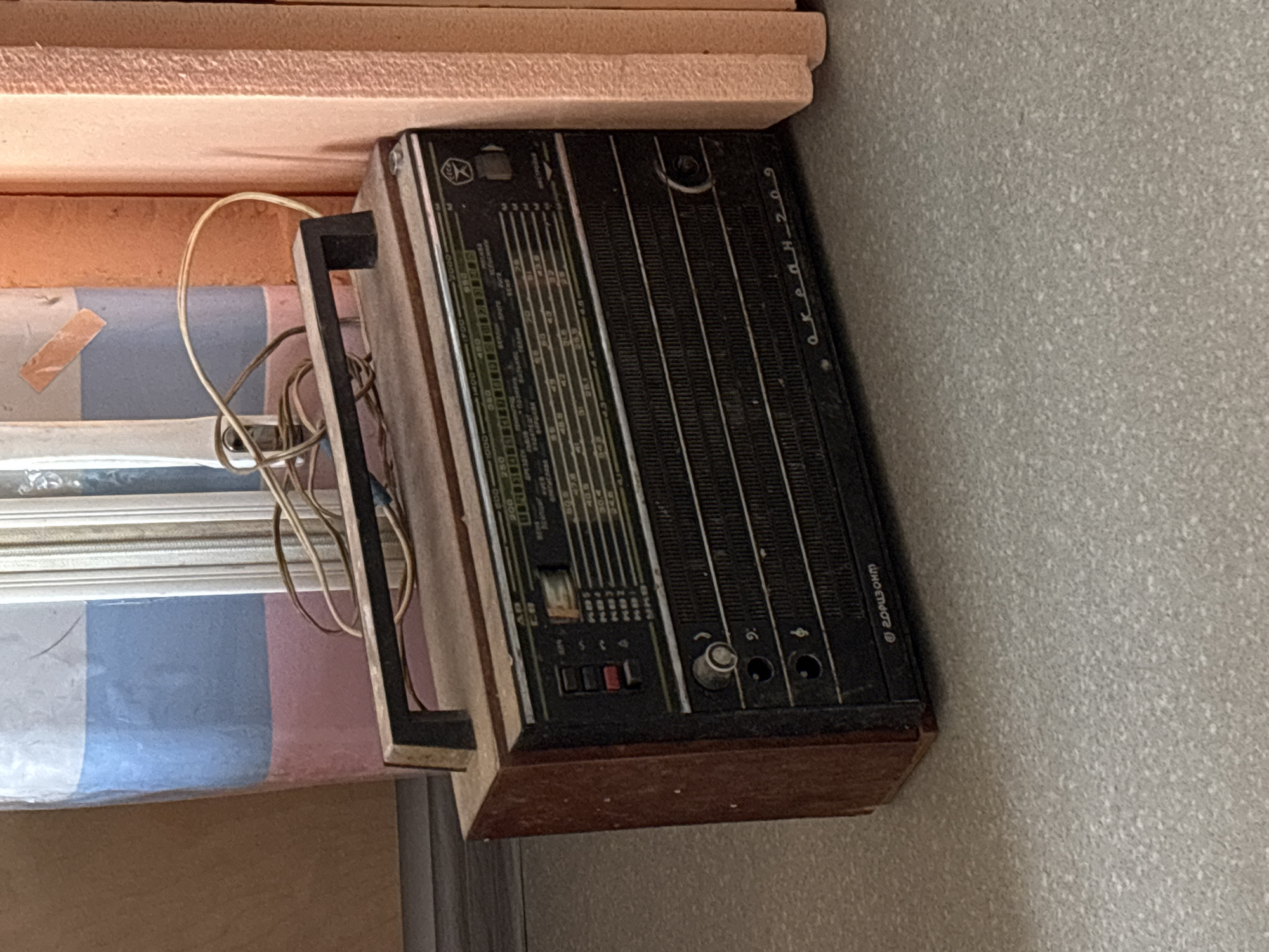A medium shot shows a vintage radio, an "Okean-209," resting on what appears to be a shelf or the edge of furniture. The radio is encased in dark wood with a black front panel featuring a tuning dial and various buttons. The inscription "Okean-209" is visible in Cyrillic script. An electrical cord is partially visible near the radio's side. The radio, manufactured by "Gorisont," is a vintage Soviet-era device. The location is not identifiable from the image.