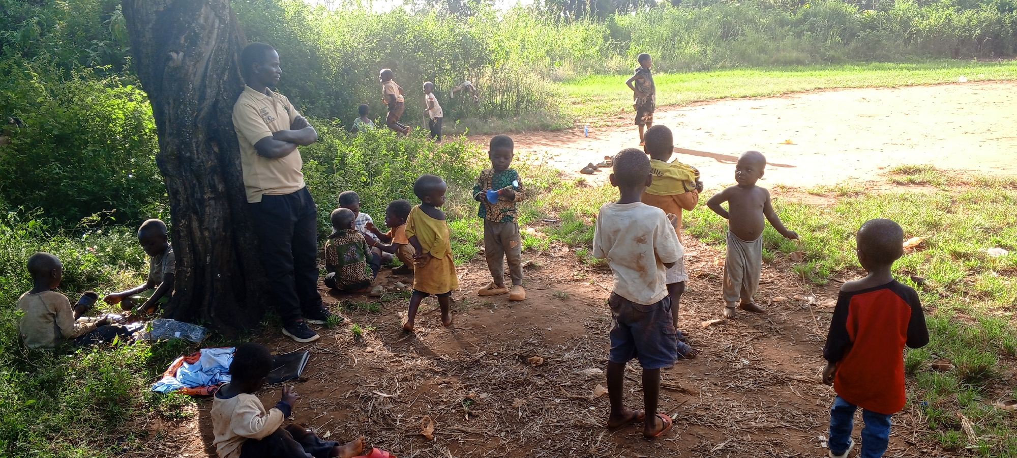 The image captures an outdoor scene in Ngalula, Democratic Republic of the Congo, featuring an adult man and numerous children gathered in a sunny, semi-rural setting. The time of day appears to be bright daytime, indicated by strong sunlight and clear shadows.
On the left side of the frame, a tall, sturdy tree trunk provides shade. Leaning against it is an adult male, wearing a light-colored polo shirt with a visible logo (which includes what looks like a 'Q' and possibly 'KPC') and dark trousers, with his arms crossed as he observes the children.
Scattered across the foreground and midground are various children, ranging from toddlers to older kids. Some are seated on the ground near the base of the tree, engaged in quiet activity, with what appear to be books or school supplies visible. One child wears a patterned shirt and holds a blue and white object, possibly a toy or bottle. Other children stand in various poses, some looking towards the background. In the distance, several children are actively playing, with one child captured mid-air, suggesting jumping or running.
The setting is characterized by a dry, reddish-brown dirt ground with patches of green grass and lush green bushes and trees forming a dense background. The ground shows some sparse dry vegetation and small pieces of litter. The children are dressed in simple, casual clothing, some barefoot and others wearing sandals. The overall scene conveys a relaxed and natural atmosphere of community and childhood play in a warm, sunny environment.