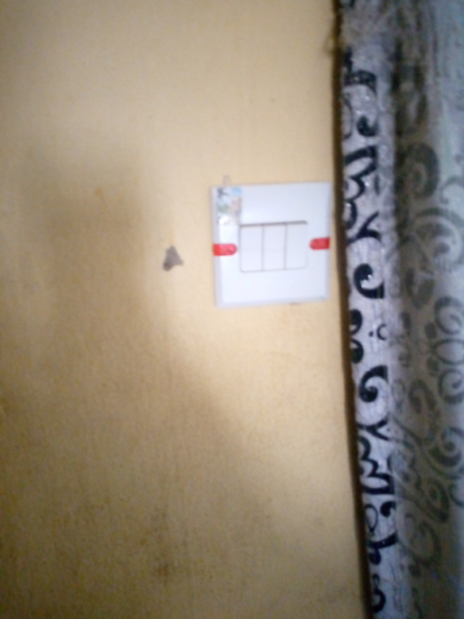 The image shows a close-up of a wall with a light switch. The wall is painted a light yellow color, and there are some subtle dark spots on its surface. To the right of the light switch, a patterned curtain is visible, with black swirls on a light gray background. The light switch itself is white and has three individual switches arranged horizontally. Red elements are visible on either side of the light switch, potentially indicating an indicator light or a decorative accent. A small, dark, triangular shape is also present on the wall to the left of the light switch. The image appears to be taken indoors, likely in a residential setting, and does not contain any discernible geographical markers to pinpoint a specific location within Jalingo, Nigeria.