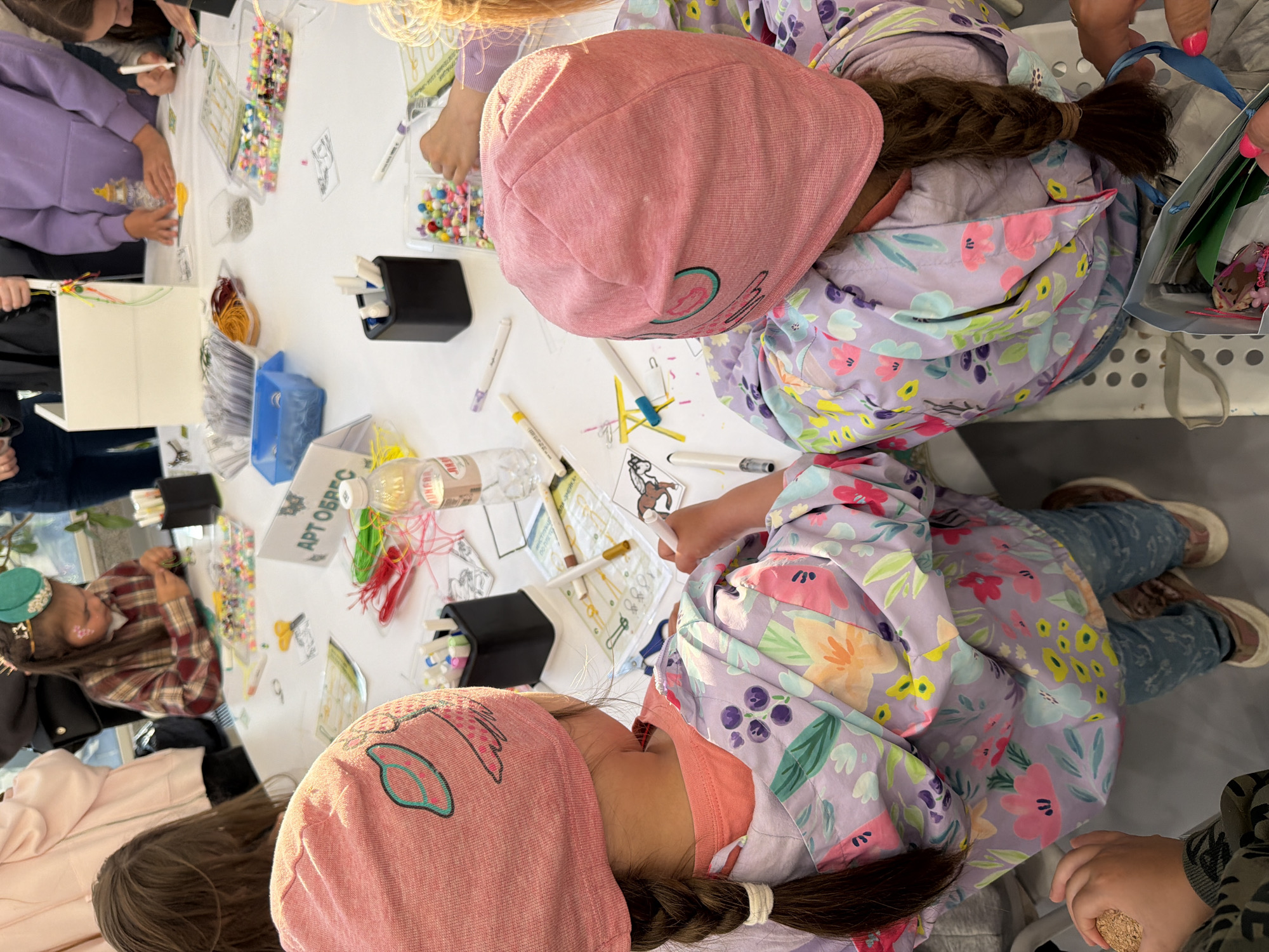 In a brightly lit, indoor setting, two young girls, facing away from the camera, are seated at a table covered with various crafting supplies. Both girls are wearing pink knit hats and floral-patterned jackets. The girl on the left has a braid of dark hair. The girl on the right has her braid visible. The table is laden with an assortment of colorful beads in clear plastic containers, spools of thread, markers, and craft paper with printed designs. One girl is holding a marker and appears to be coloring a design of a horse. A bottle of water and a small black container holding pens are also visible on the table. The background shows other people, suggesting a group activity or a public event. The overall scene depicts a children's craft workshop.