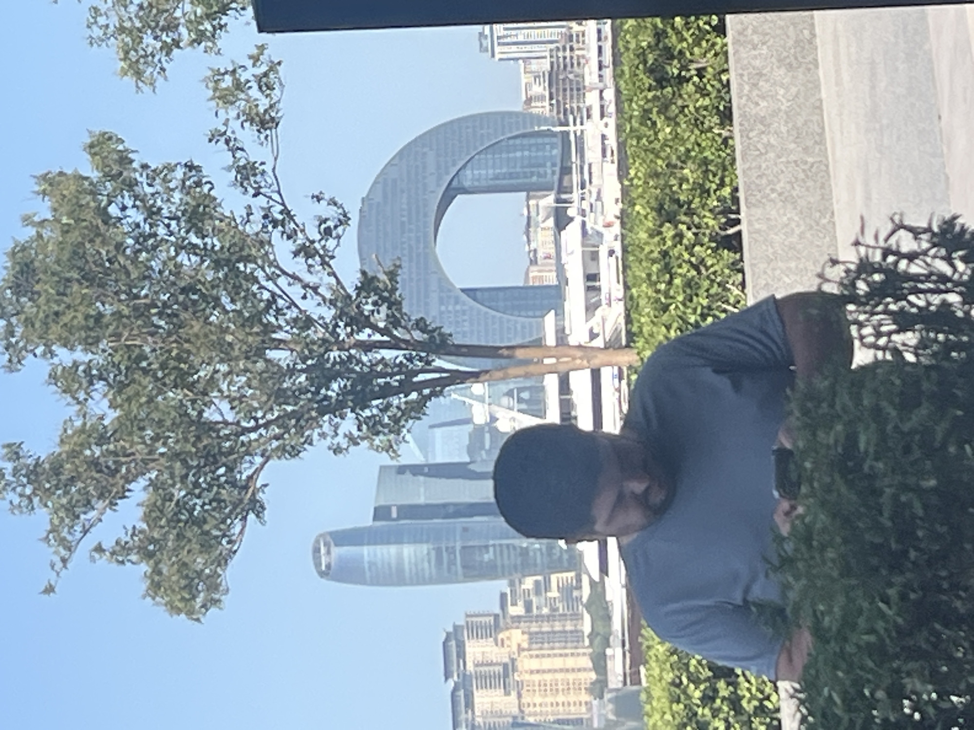 A man in a blue t-shirt is looking down at his phone, partially obscured by a green bush. In the background, the Flame Towers, a trio of skyscrapers in Baku, Azerbaijan, are visible against a clear blue sky. To the right of the Flame Towers is the Crescent Bay development, a distinctive arched building. Other modern buildings and yachts docked in the harbor can also be seen. The scene appears to be set outdoors, possibly in a park or public space with landscaping.