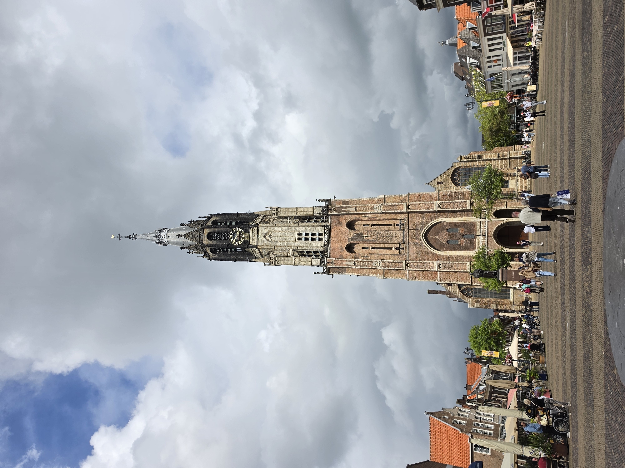 The image shows a wide shot of the Markt square in Delft, Netherlands, dominated by the towering Nieuwe Kerk (New Church). The church, made of brick with gothic architectural elements, has a tall, slender spire topped with a weather vane. The clock face is visible on the tower. The sky above is dramatic, with a mix of dark, heavy clouds and patches of bright blue sky.
The Markt square is paved with patterned bricks and is bustling with activity. Several people are walking across the square, some in groups, others individually. Some are dressed casually, suggesting it is daytime. There are outdoor seating areas for cafes and restaurants lining the square, with tables and umbrellas set up. People are seated at some of these tables.
On the left side of the square, there are buildings with traditional Dutch architecture, featuring gabled roofs and orange tiles. A statue of a man is visible in front of the church. Bicycles are parked in various locations around the square. The overall impression is a vibrant public space in a historic European city.