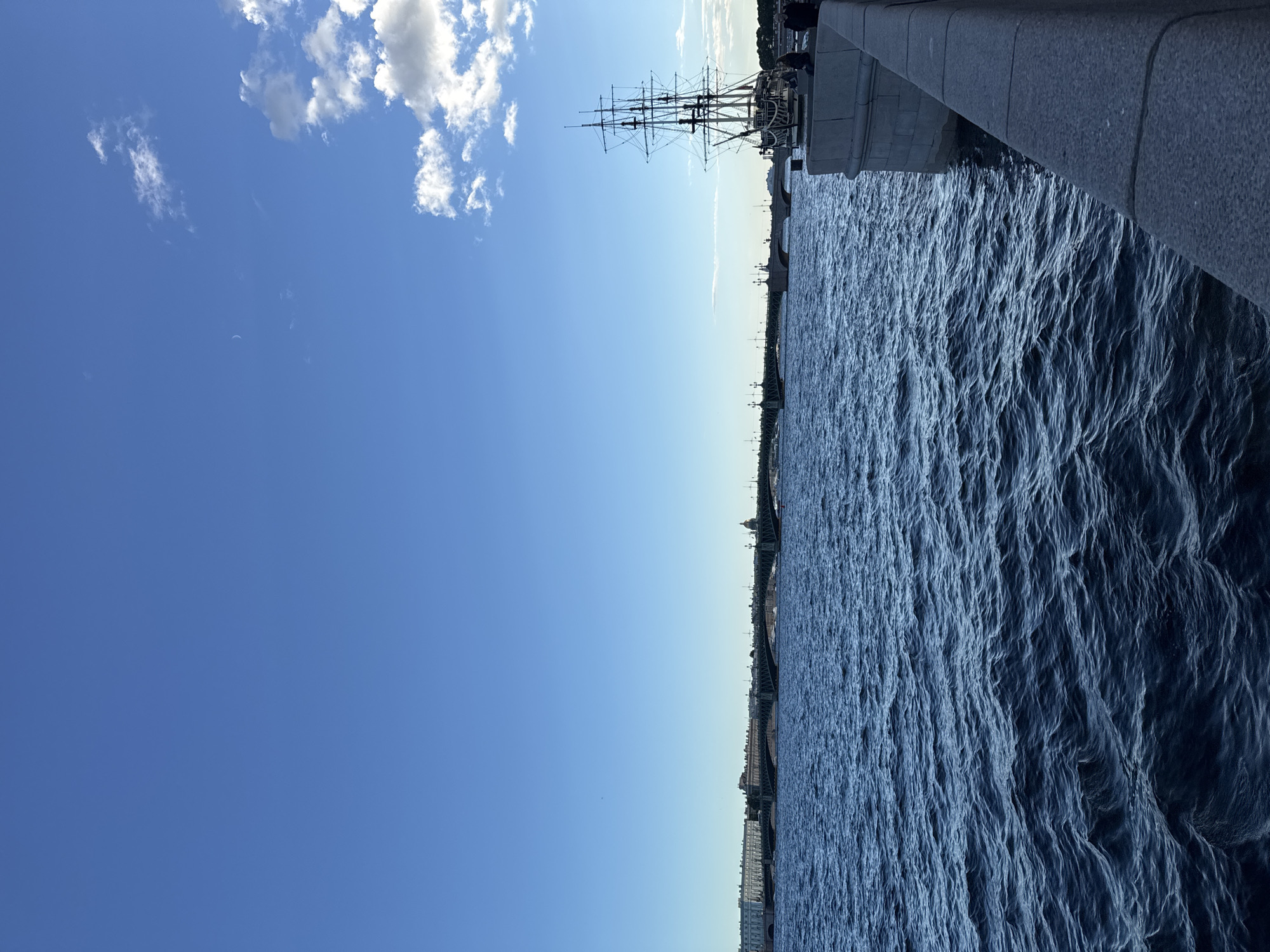 An image depicts a serene scene along a wide river in Saint Petersburg, Russia, under a clear blue sky with scattered white clouds. In the foreground, the dark blue water of the river is rippled, suggesting a gentle breeze. Along the right bank, a stone embankment curves into the distance, topped by a large, ornate sailing ship with multiple masts and rigging. Several people are visible on the embankment, some seated and others standing near the ship. Across the river, a long, multi-arched bridge spans the water, connecting the city's districts. Buildings with a classical architectural style line the far bank, indicating the historic center of Saint Petersburg. The overall impression is one of calm and historic grandeur.