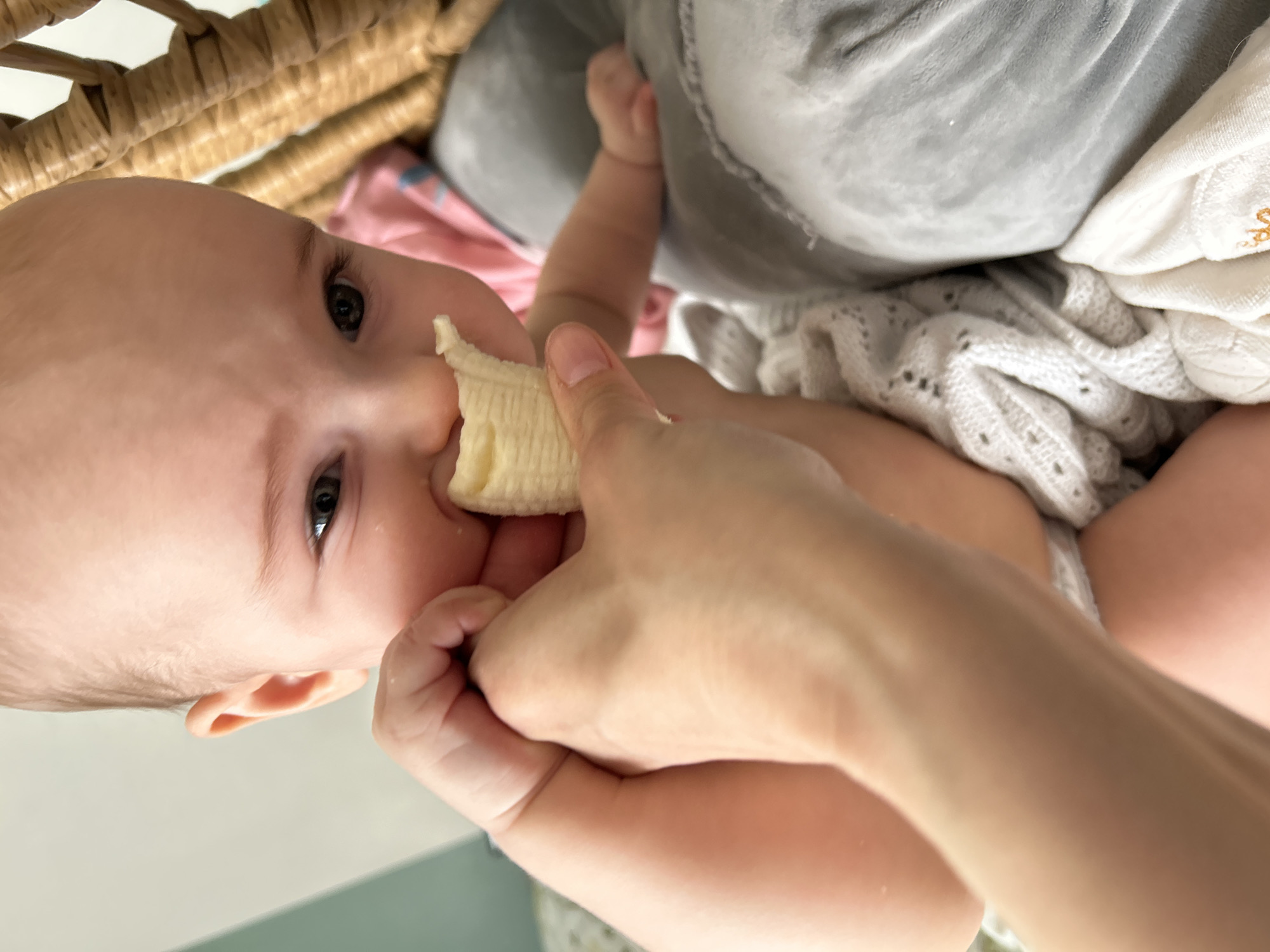A baby is being fed a piece of banana by a person's hand. The baby is lying down, partially covered by a white knitted blanket and other soft materials. A woven basket is visible in the background. The location is not discernible as being in Moscow, Russia.