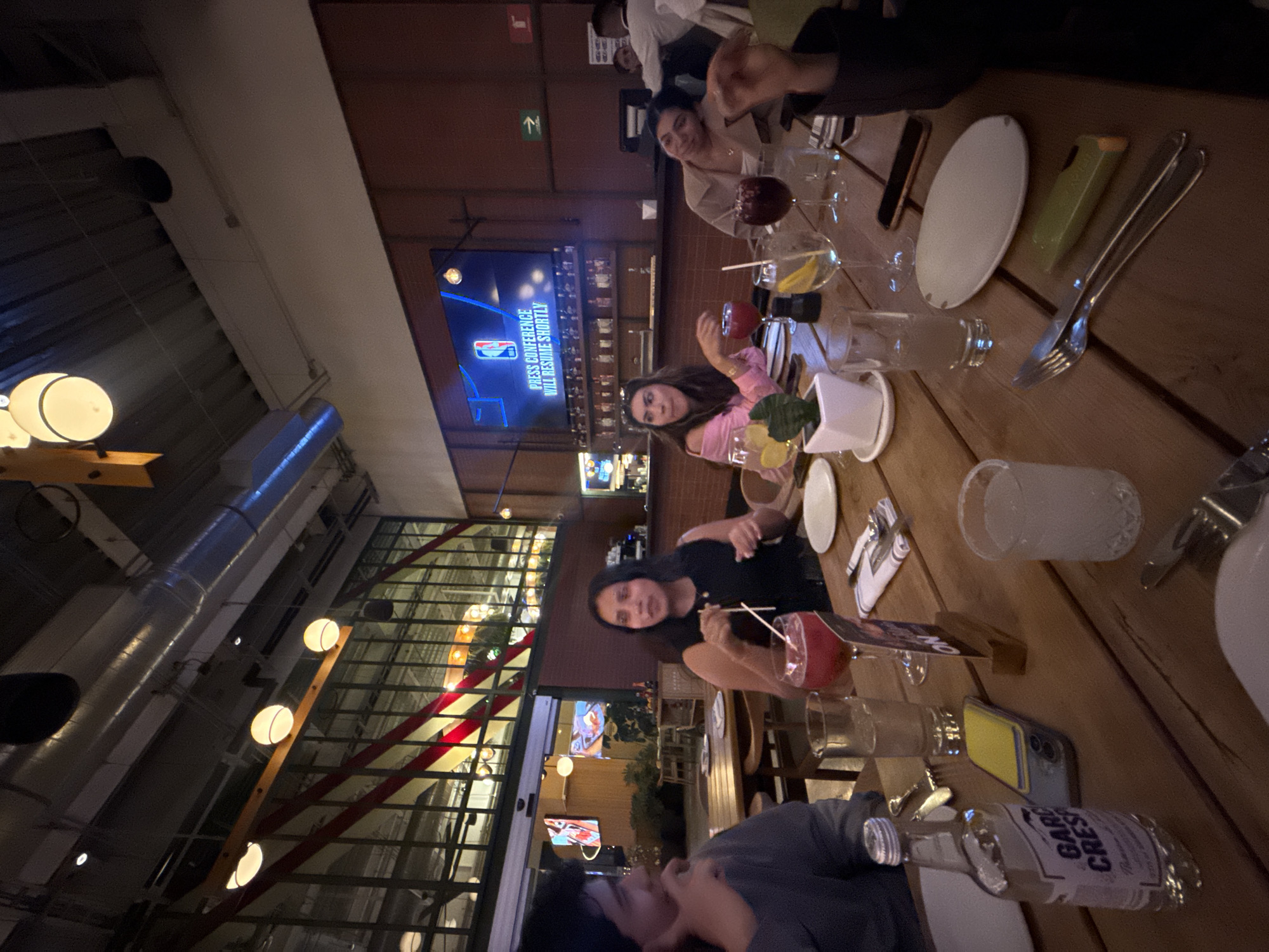 The image shows a group of people gathered around a wooden table at a restaurant in Mexico City, Mexico. Three women and one person are visible. The setting is a dimly lit indoor space with large windows. The table is covered with plates, glasses, and drinks. The restaurant's design features industrial elements, like metal pipes and exposed bulbs. There's a large TV screen in the background displaying NBA programming.