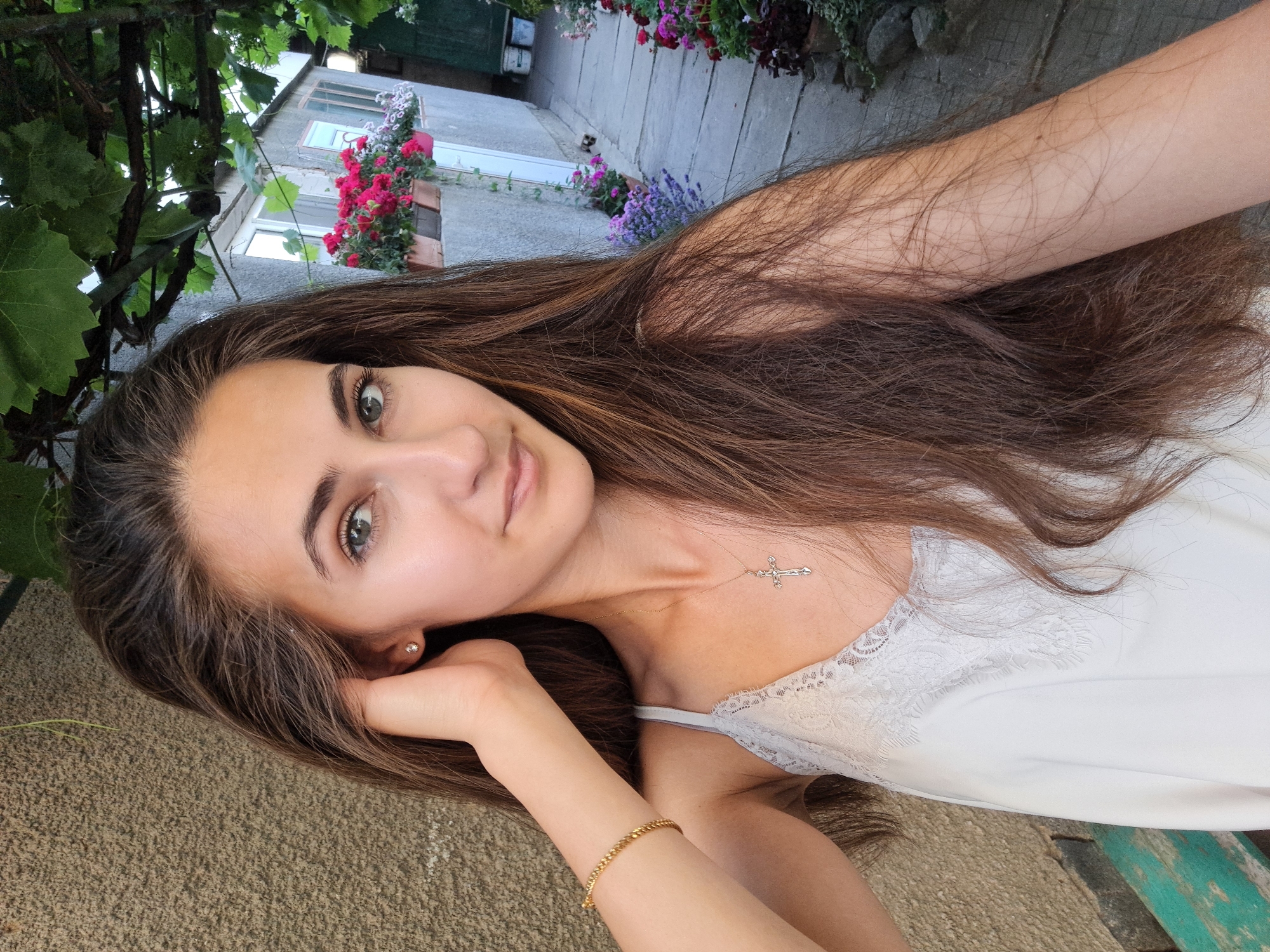 A young woman with long brown hair is taking a selfie outdoors, in what appears to be a rural or semi-rural setting. She is wearing a white lace-trimmed camisole and a gold necklace with a cross pendant. A gold bracelet is visible on her left wrist. Her left hand is resting on her head.
Behind her, to the left, is a textured, beige wall. To the right, a stone pathway leads towards a small, weathered building with windows. Potted flowers, including pink and white varieties, are displayed along the building's exterior. Lush green leaves from an overhead vine are visible in the upper part of the image. The presence of the building, pathway, and potted plants suggests a residential yard or garden. The building's architectural style, the plants, and the overall atmosphere are consistent with a location in Chepintsi, Bulgaria. The lighting suggests it is daytime.