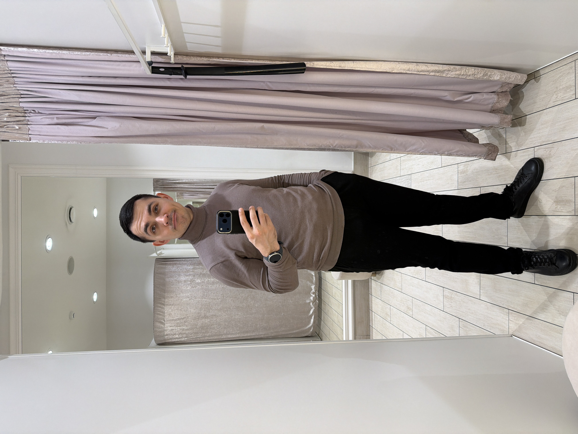 A man is taking a selfie in what appears to be a dressing room or changing room. He is wearing a brown turtleneck sweater, black pants, and black shoes. He is holding a black smartphone in his left hand, with his wrist visible, displaying a watch. The changing room has a light-colored curtain hanging from a rod, and the floor is tiled. The walls are painted white. The man's face is visible in the reflection of a mirror. It is not possible to determine if the image was taken in the city of Izhevsk, Russia.