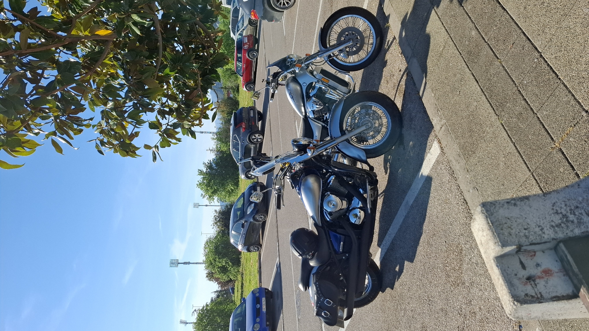 Two motorcycles are parked on a paved parking lot. In the background are several cars. Trees and light poles are also visible. The location is assumed to be in Bar, Montenegro.