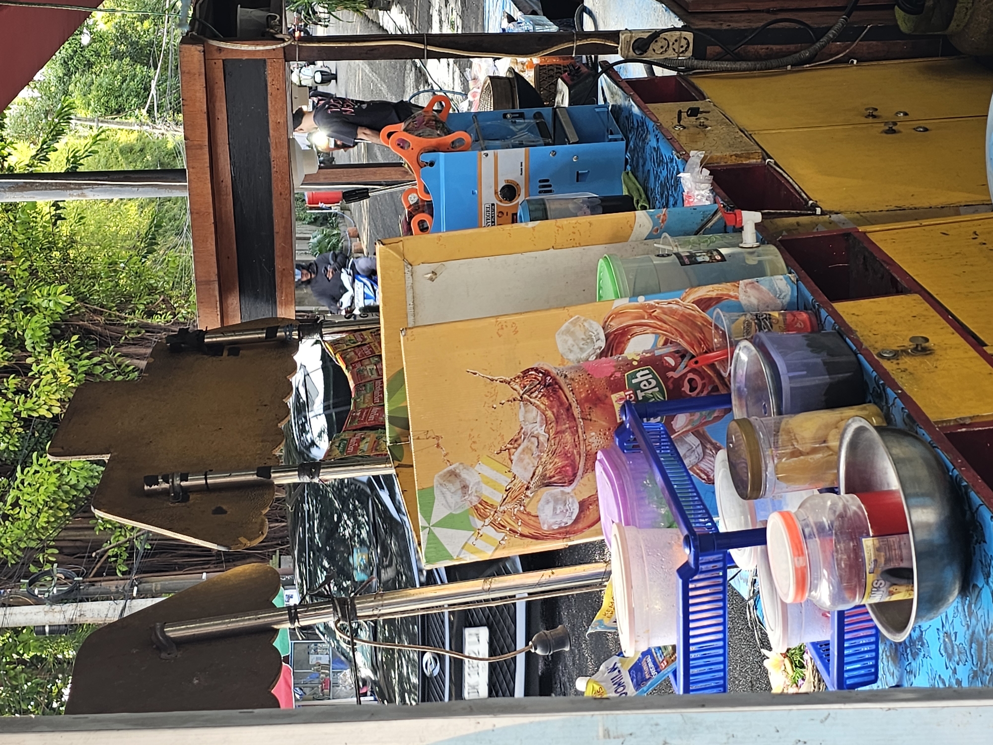 The image shows a street food vendor's cart in Denpasar, Indonesia. The cart is laden with containers of ingredients, including liquids, powders, and solids, along with associated serving items like cups and bowls. The cart has yellow and blue elements. A person in a dark shirt and white hat is seen near a blue machine on the cart. Outside of the cart, other people are visible walking along the street. Foliage is visible beyond the cart.