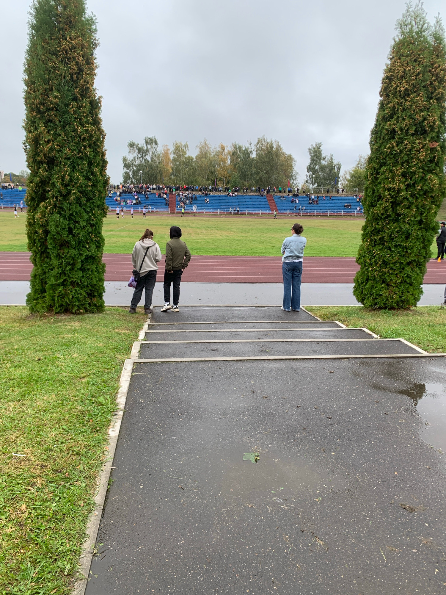 The image depicts an outdoor track and field stadium on an overcast day. In the foreground, a wet asphalt path leads up a set of stairs. Two individuals, a person wearing a grey hoodie and another in a dark jacket, stand on the grass to the left, facing away from the camera. To the right, a person in a denim jacket and blue jeans stands on the asphalt, also facing away. Beyond them, a stadium with blue bleachers filled with spectators overlooks a green field. Several trees flank the stadium, with two tall, dense evergreen trees on either side of the frame. The overall atmosphere suggests a sporting event in progress or about to commence in Kapyĺ, Belarus.