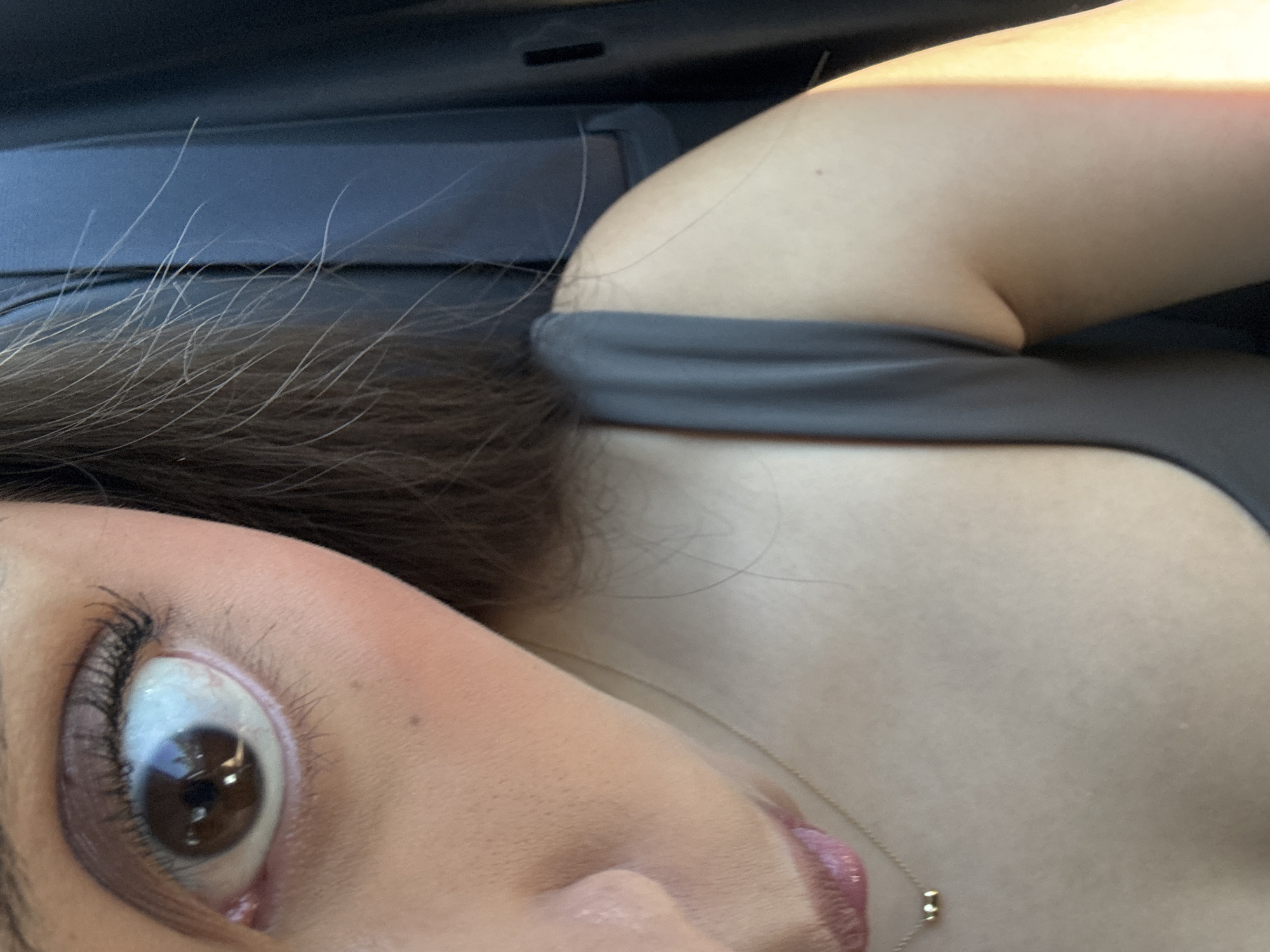 A close-up, high-angle portrait shows a person partially visible, likely taking a selfie inside a vehicle. The subject’s right eye, nose, mouth, and a portion of their chest and shoulder are in the frame, tilted slightly. The eye is wide open, with a rich brown iris reflecting bright light, possibly sunlight, and long, dark eyelashes. The person has fair skin, with visible pores and a couple of small moles on the cheek and shoulder. Pink lip gloss or lipstick is applied to the lips. A delicate gold chain necklace with three small, circular elements hangs around the neck. The person is wearing a dark, possibly grey, top or strap. Long, dark hair frames the face, with some lighter strands visible due to lighting. The background consists of dark grey and blue tones, consistent with a car interior, possibly a seat or overhead compartment. Bright illumination suggests daytime. The image captures a casual moment in San Bernardino Tlaxcalancingo, Mexico. No visible text is present.