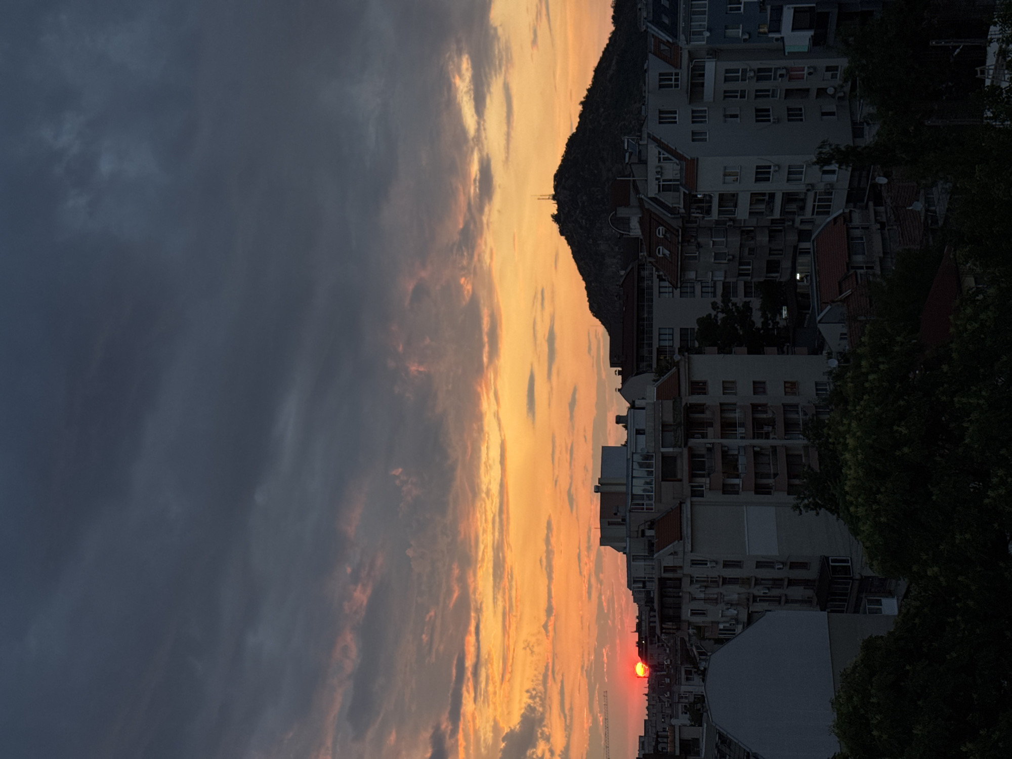 The image shows a sunset over the city of Plovdiv, Bulgaria. The sun, a bright red orb, is setting behind a hill that has a telecommunications tower on its summit, a prominent landmark in Plovdiv known as Bunardzhik Hill or Alyosha Hill. The sky is a dramatic mix of dark gray clouds, tinged with orange and pink hues from the setting sun, creating a vibrant contrast. Below the sky, the silhouette of the city's buildings is visible. The buildings are a mix of architectural styles, with some appearing to be older residential blocks and others more modern structures. The foreground is dominated by lush, dark green trees, obscuring the lower parts of the buildings and adding depth to the scene. The overall impression is a city bathed in the warm, fading light of sunset, with the iconic hill serving as a backdrop.